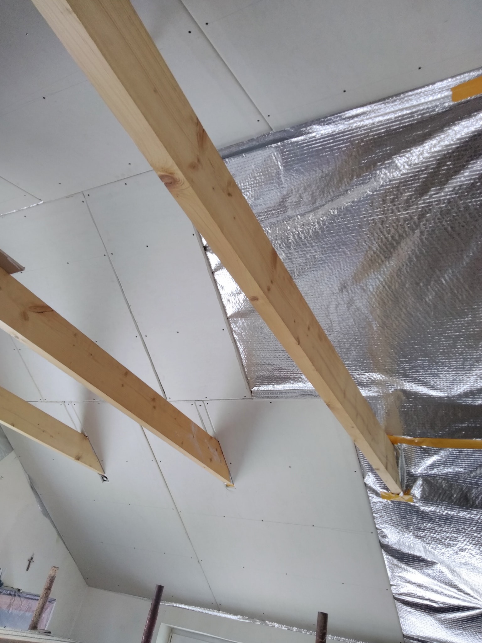 Skośny sufit poddasza w trakcie prac: widoczne drewniane belki konstrukcyjne, płyty gipsowo-kartonowe oraz warstwa izolacji termicznej z folią aluminiową.