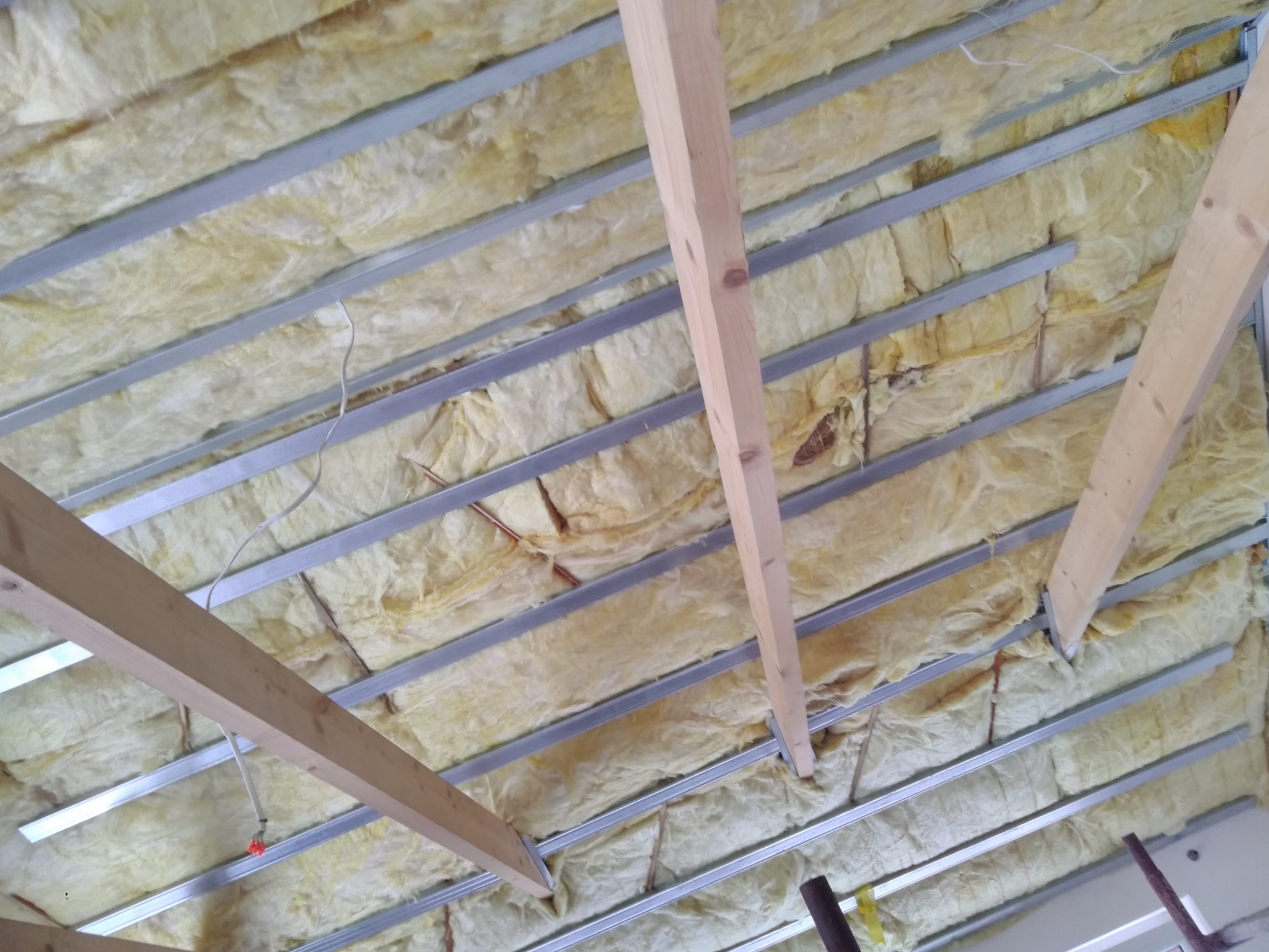 Izolacja poddasza wełną mineralną, widoczne metalowe profile i drewniane elementy konstrukcyjne, przewody elektryczne w tle.