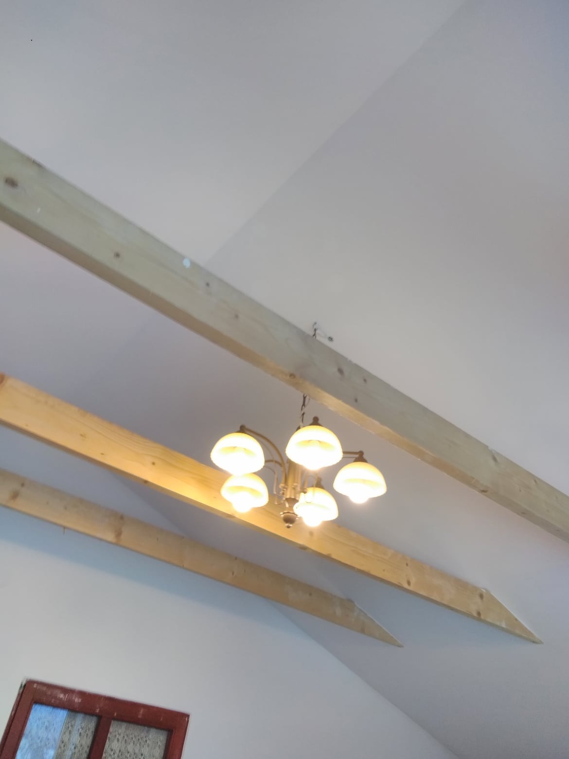 Poddasze z widocznymi drewnianymi belkami konstrukcyjnymi i wiszącą lampą z pięcioma mlecznymi kloszami, częściowo widoczne okno w czerwonej ramie.