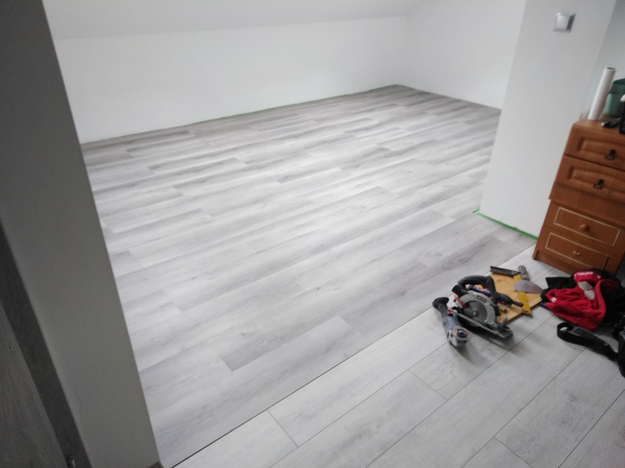 Jasnoszara podłoga panelowa ułożona w pokoju na poddaszu, widoczne narzędzia do cięcia paneli oraz komoda w rogu pomieszczenia.