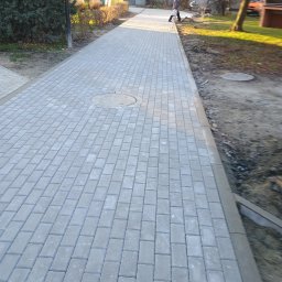 BRUKO-BUD MATEUSZ KOBYLSKI - Nowo ułożony chodnik z szarej kostki brukowej, widoczny właz kanalizacyjny, obrzeża chodnika i fragment terenu z przygotowanym podłożem, w tle budynki mieszkalne i przechodząca osoba.