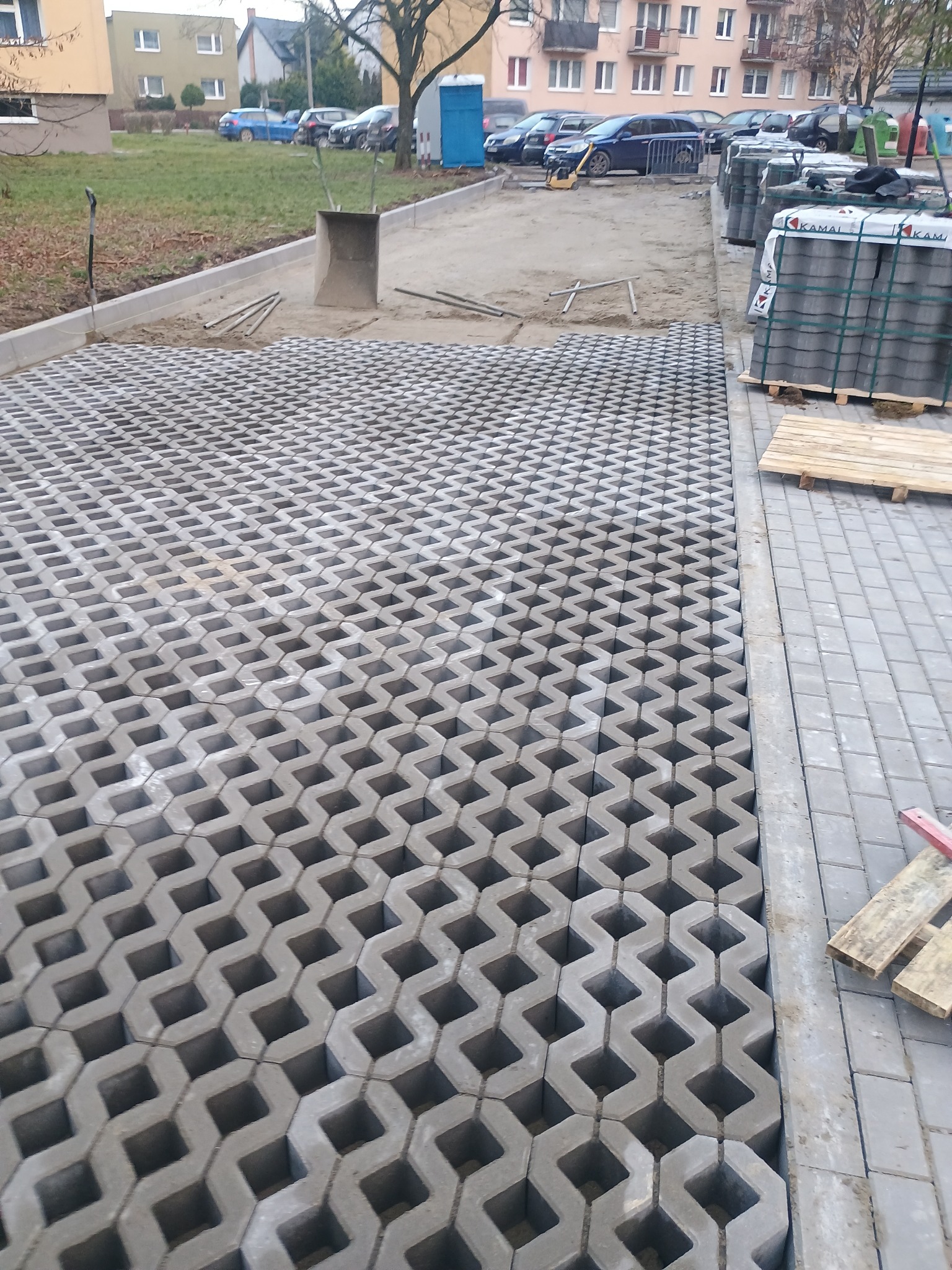 Układanie ażurowych płyt betonowych na przygotowanym podłożu, widoczne palety z materiałem i ubijarka w tle, teren częściowo utwardzony kostką.