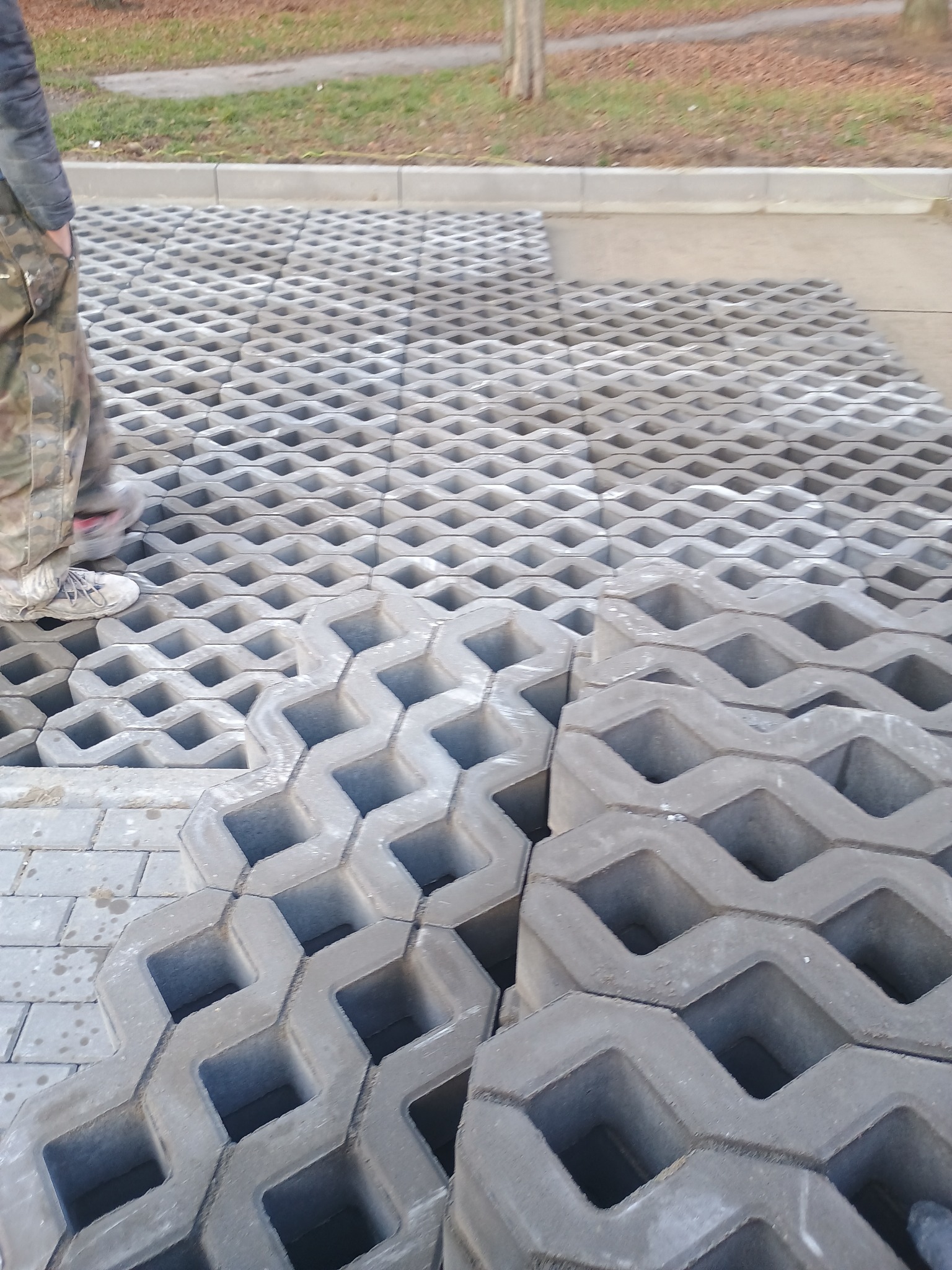 Układanie ażurowych płyt betonowych na podjeździe, widoczne ułożone płyty i stos płyt do ułożenia, częściowo widoczna osoba w ubraniu roboczym.