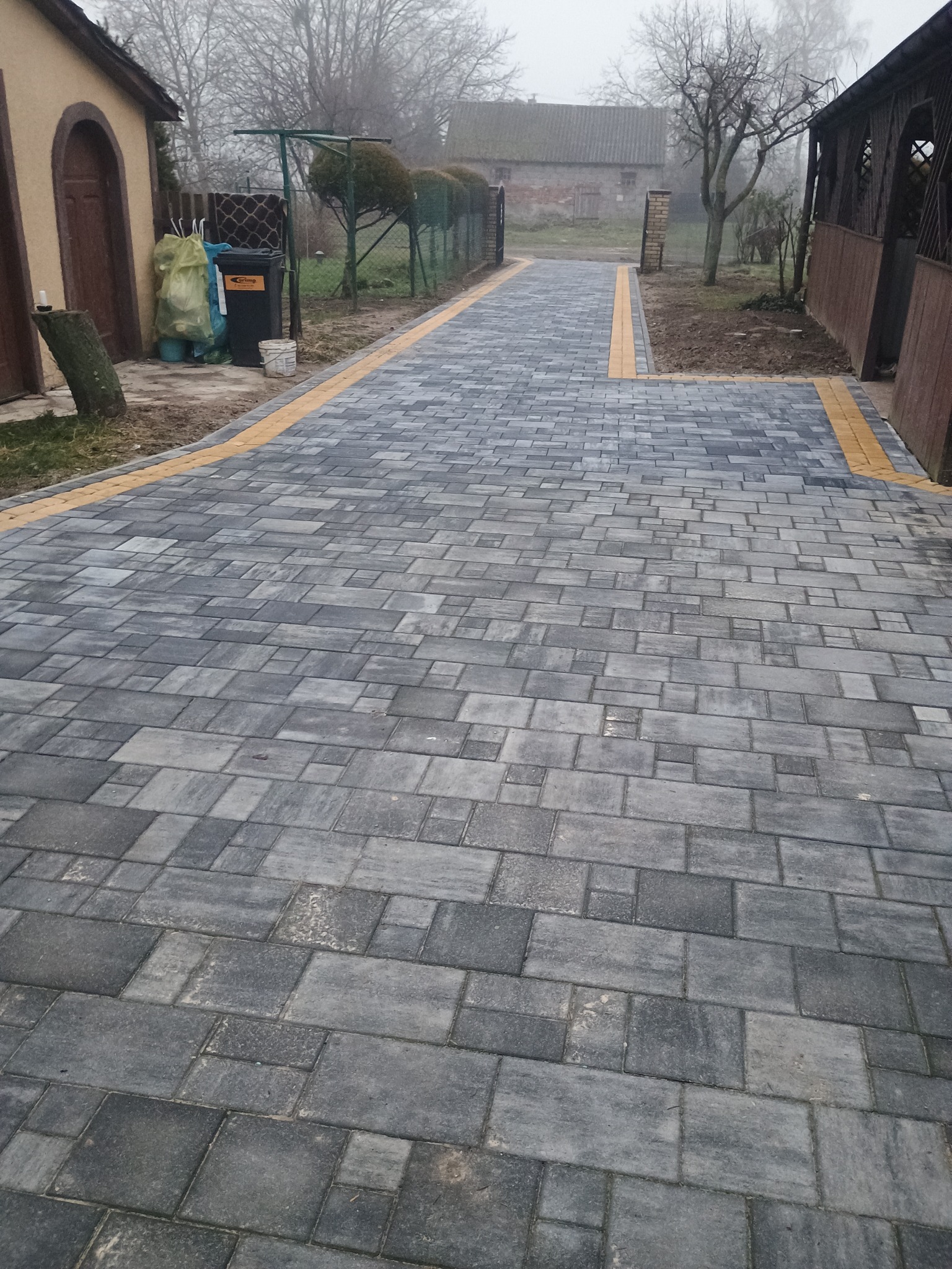 Ułożona ścieżka z szarej kostki brukowej z żółtymi obrzeżami, prowadząca przez posesję w mglisty dzień.