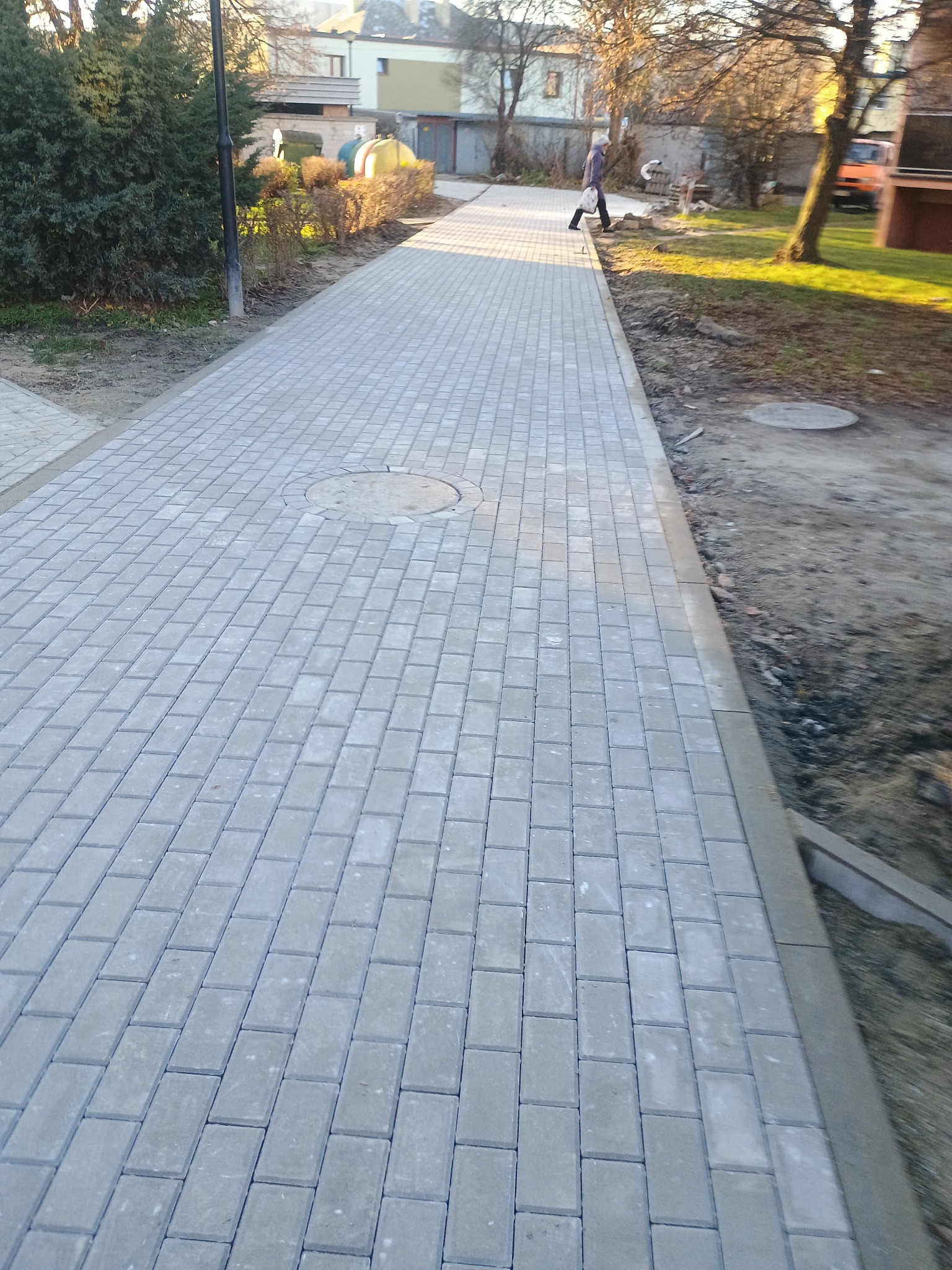 Nowo ułożony chodnik z szarej kostki brukowej, widoczny właz kanalizacyjny, obrzeża chodnika i fragment terenu z przygotowanym podłożem, w tle budynki mieszkalne i przechodząca osoba.