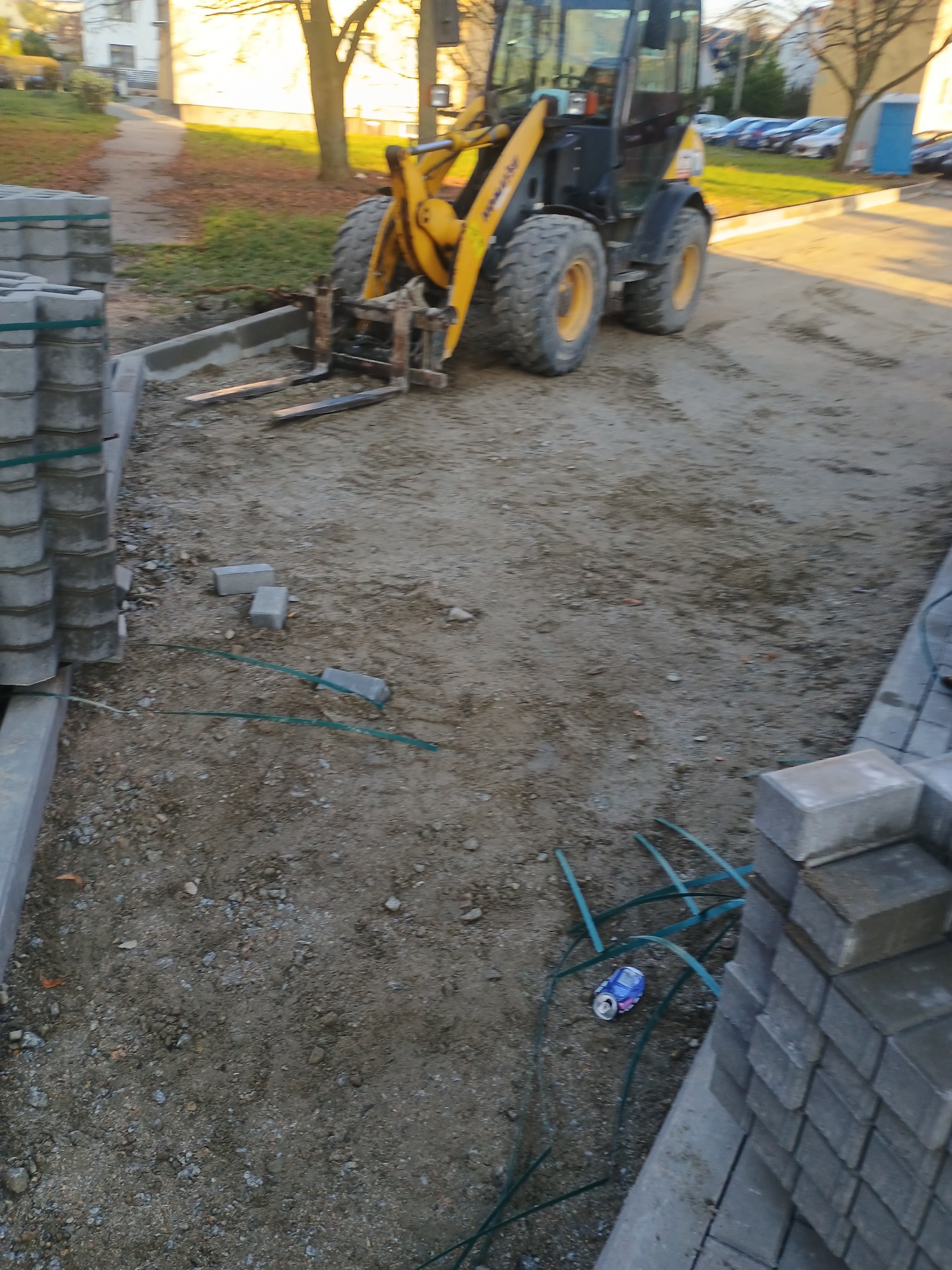 Żółta ładowarka teleskopowa na placu budowy, obok palety szarych kostek brukowych i krawężników, widoczna puszka po napoju