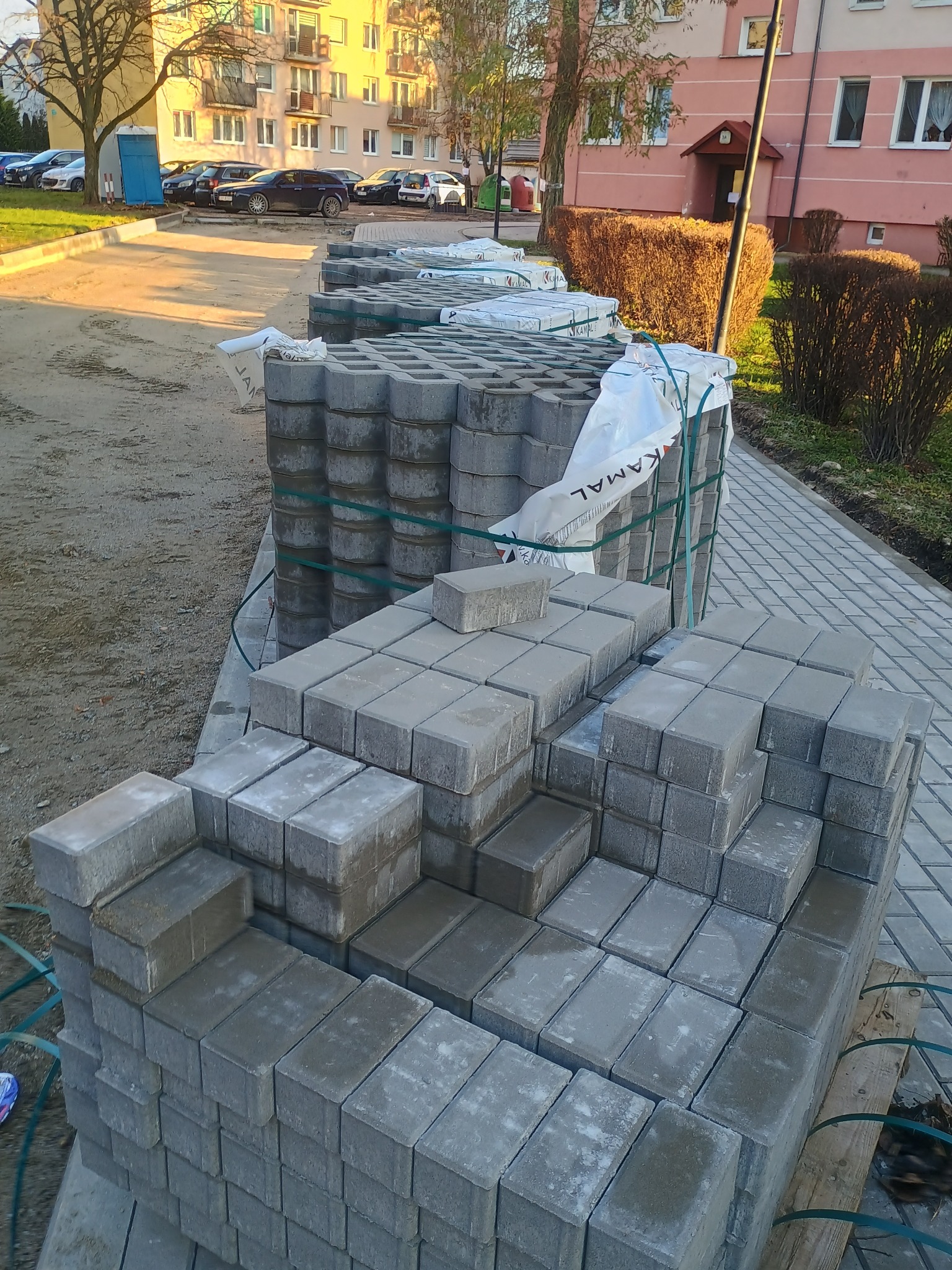 Szare betonowe kostki ułożone na paletach, czekające na ułożenie, z widocznym fragmentem już ułożonej nawierzchni i budynkami w tle.