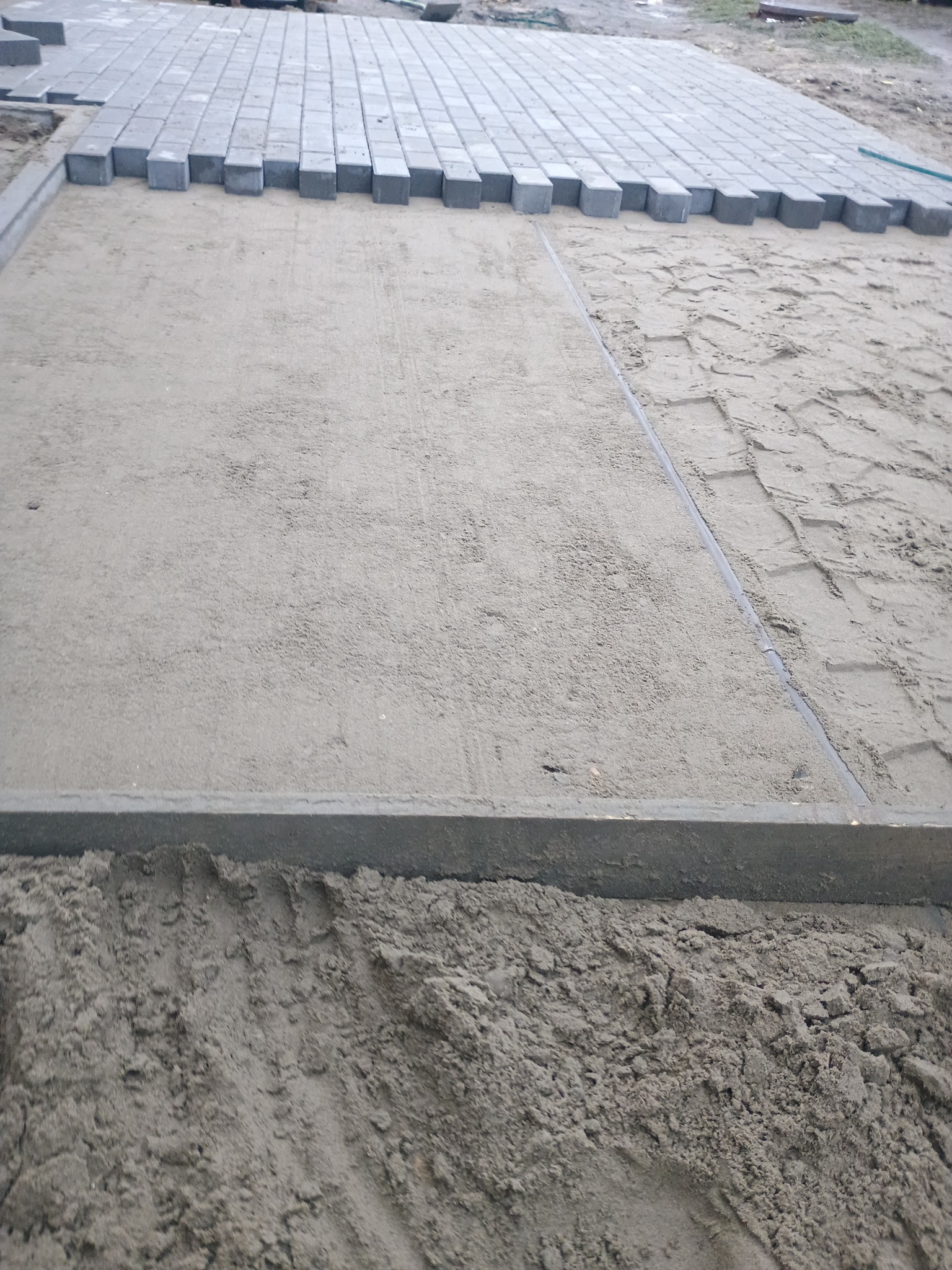 Układanie szarej kostki brukowej na podsypce piaskowej, widoczne krawężniki obramowujące teren.