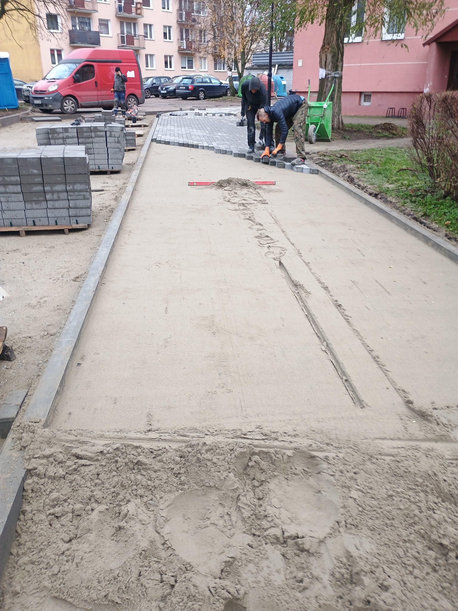 Prace brukarskie w toku: przygotowane podłoże z piasku, częściowo ułożona szara kostka, robotnicy przy pracy, paleta z kostką w tle, budynki mieszkalne w tle.