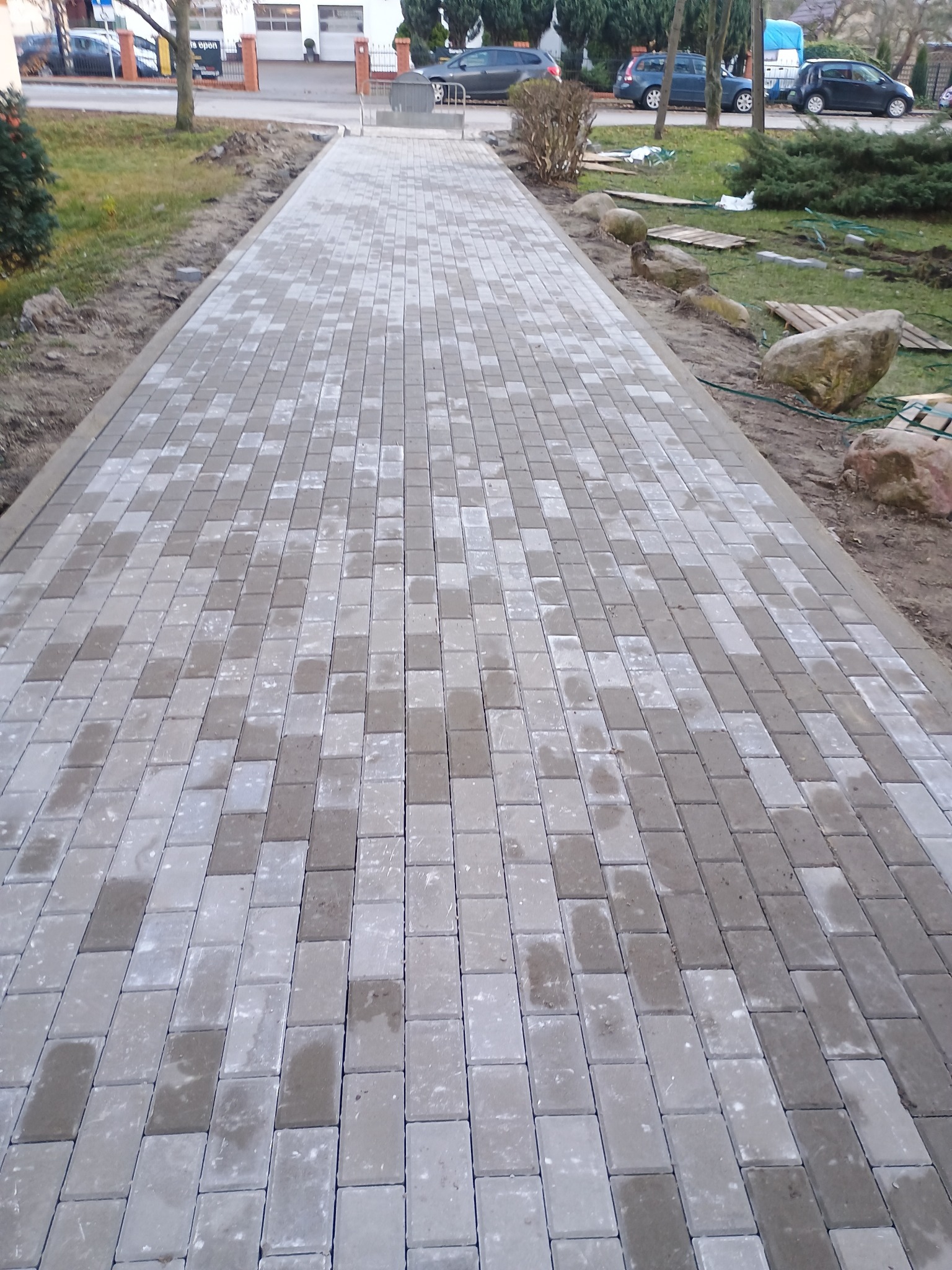 Ułożona ścieżka z prostokątnej, szarej kostki brukowej, widziana z perspektywy osoby idącej, z widocznym trawnikiem po bokach, na którym leżą kamienie i drewniane palety.