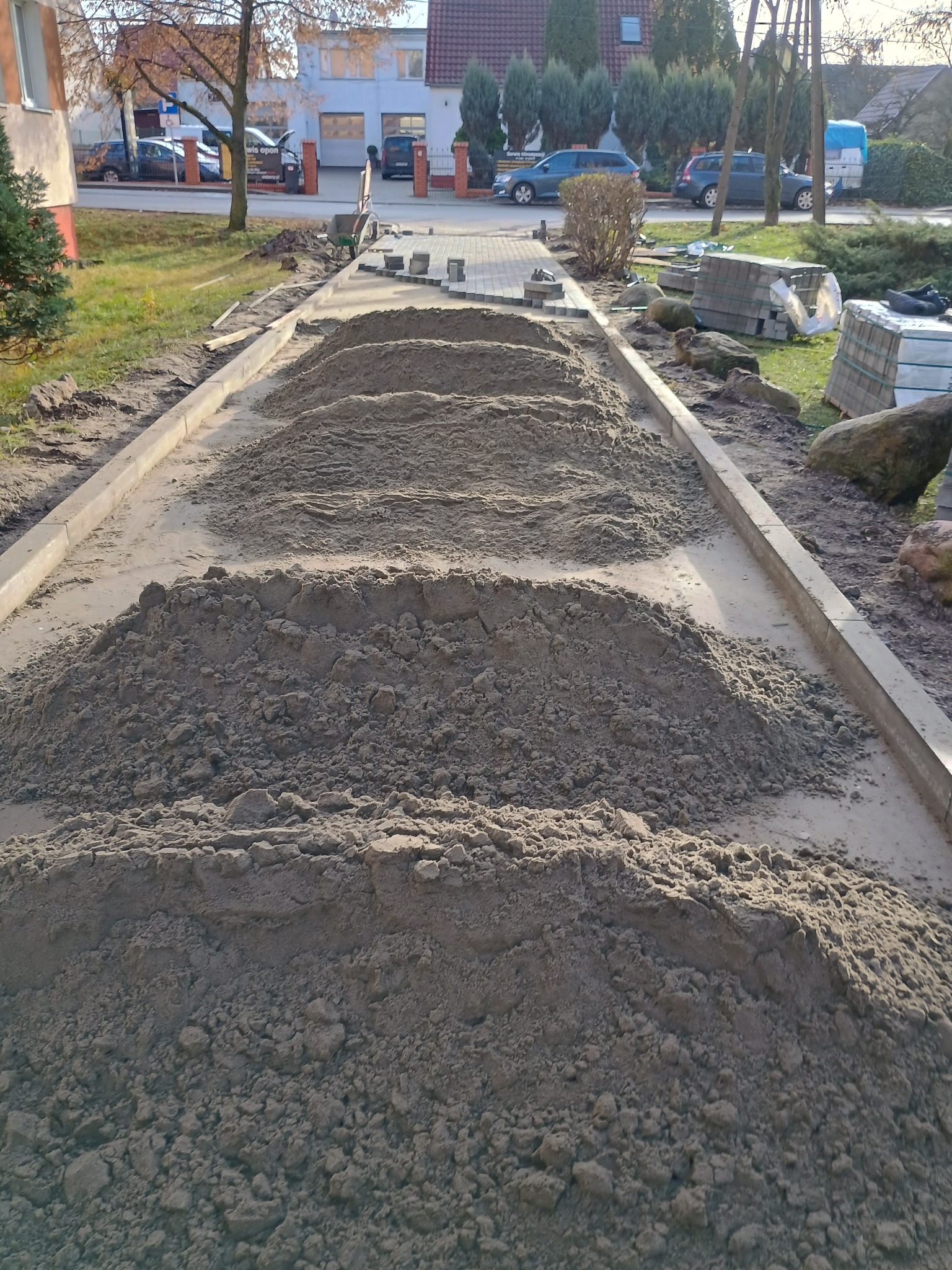 Przygotowanie podłoża pod ścieżkę z ułożonymi obrzeżami; widoczne pryzmy piasku, kostka brukowa w tle i budynki mieszkalne.