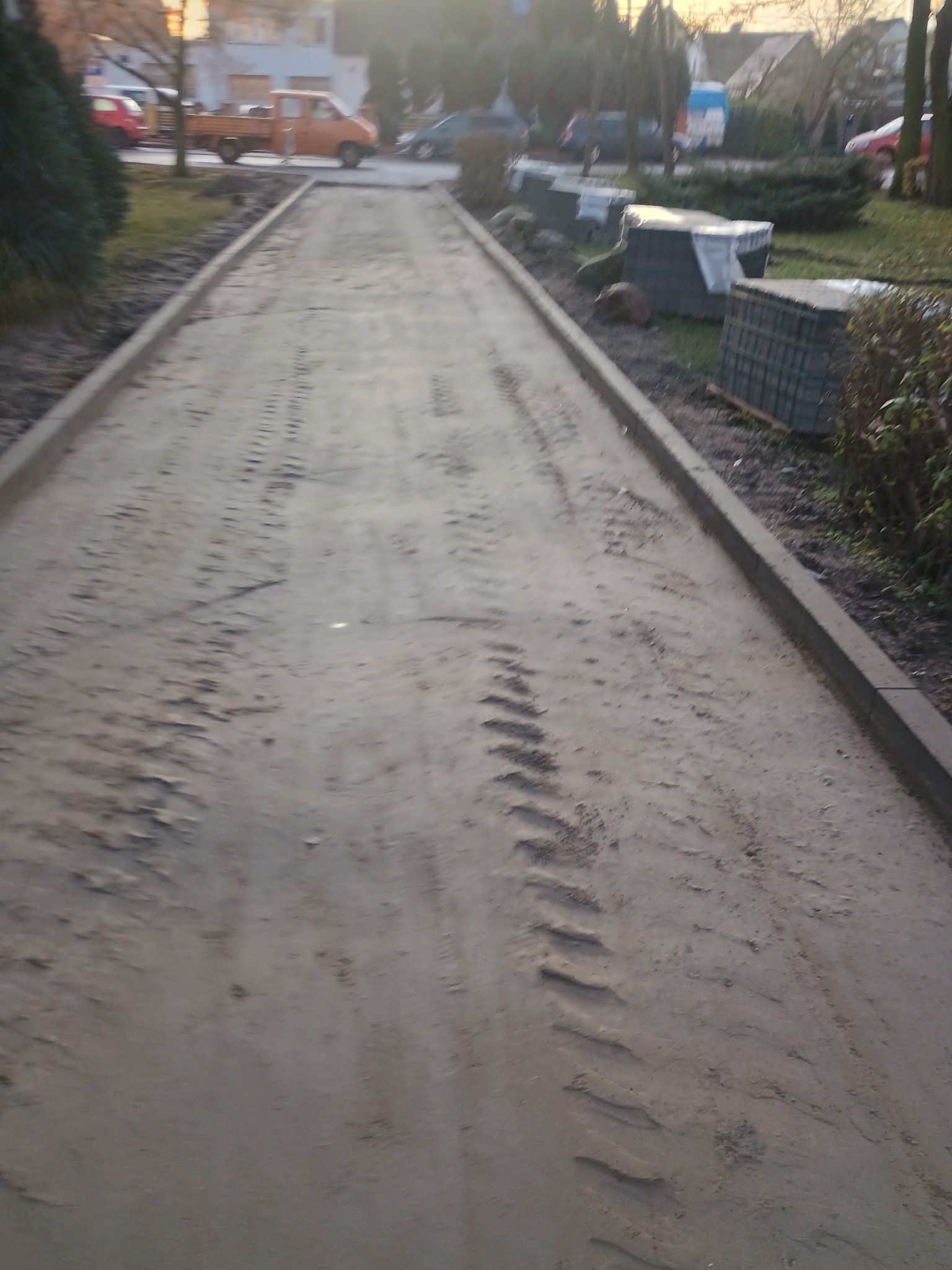 Przygotowany teren pod budowę drogi z widocznymi śladami opon, obrzeża chodnikowe po bokach, w tle zaparkowany pomarańczowy samochód dostawczy i stosy kostki brukowej.