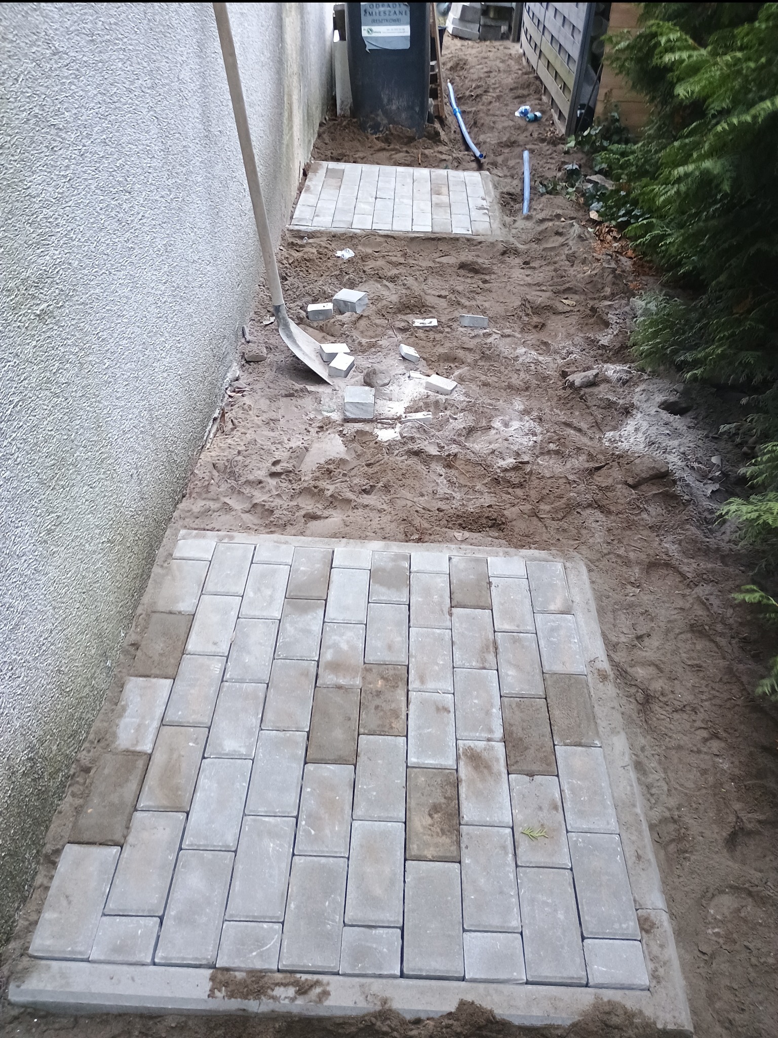 Fragment ścieżki z szarych kostek ułożonej w ogrodzie, widoczna łopata oparta o ścianę budynku i rozsypane kostki w tle.