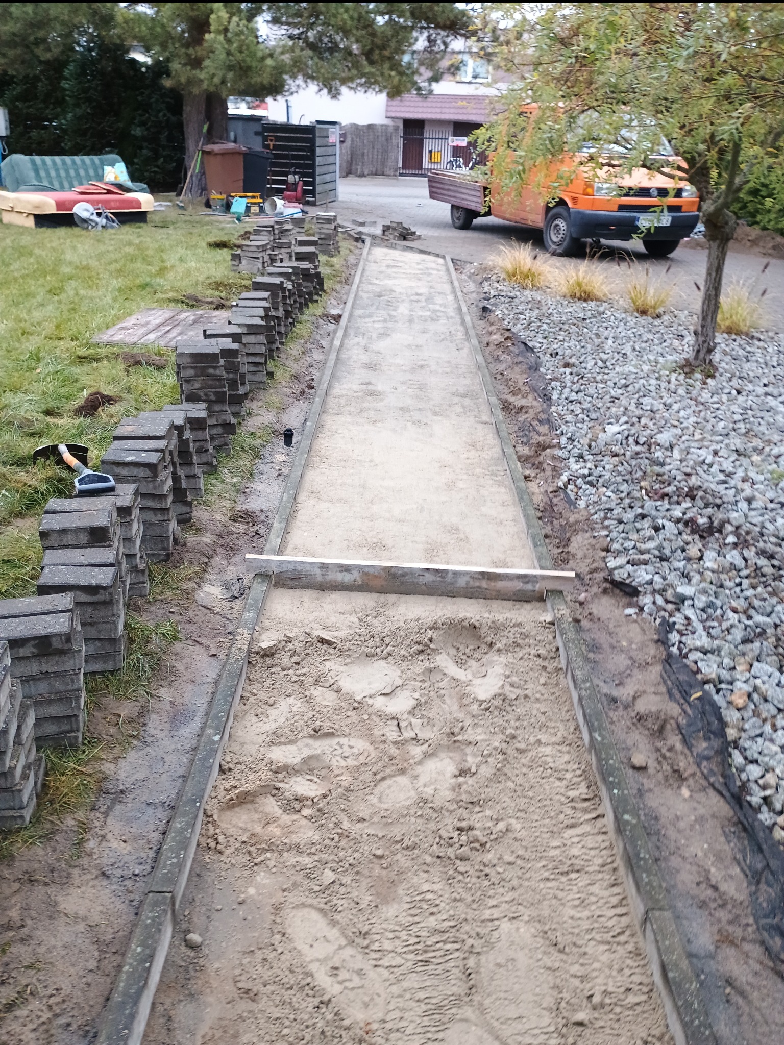 Przygotowanie podłoża pod ścieżkę z kostki brukowej, widoczne ułożone obrzeża, piasek, poziomica i stosy kostki brukowej, w tle pomarańczowy samochód dostawczy.