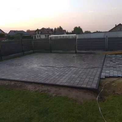 Ukończona powierzchnia z kostki brukowej w Świeciu, otoczona trawnikiem i ogrodzeniem panelowym, widok na domy w oddali podczas zachodu słońca.