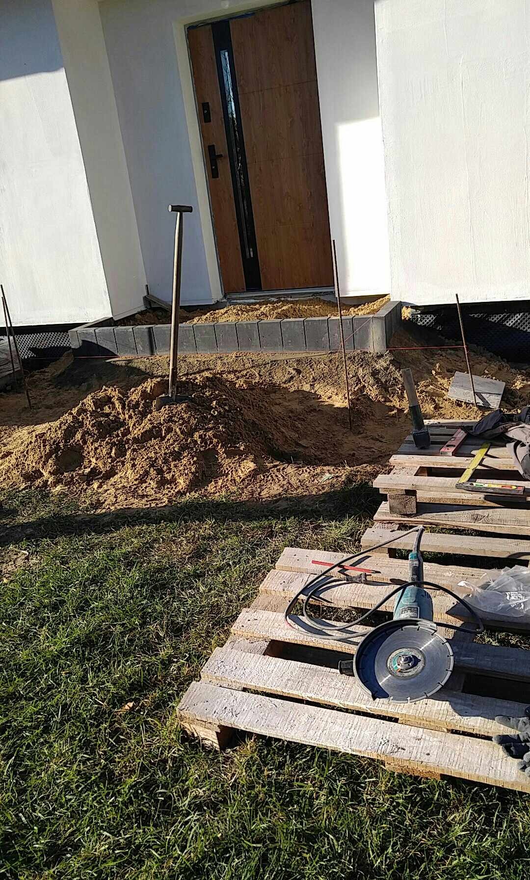 Przygotowanie terenu przed wejściem do domu: ułożone obrzeża z kostki, pryzma piasku, wbita łopata, paleta z narzędziami, w tym szlifierka kątowa z tarczą diamentową do cięcia.