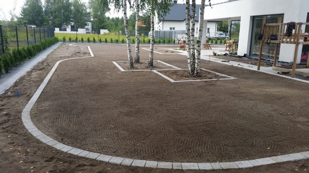 Przygotowany teren pod trawnik z obrzeżami z kostki i wyznaczonymi kwadratami wokół drzew, widoczne ślady po maszynach przygotowujących podłoże.