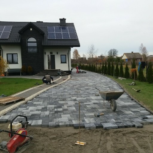 Układanie szarej kostki brukowej na podjeździe przed domem z panelami słonecznymi, widoczny pracownik doglądający ułożenia, taczka i zagęszczarka w tle.