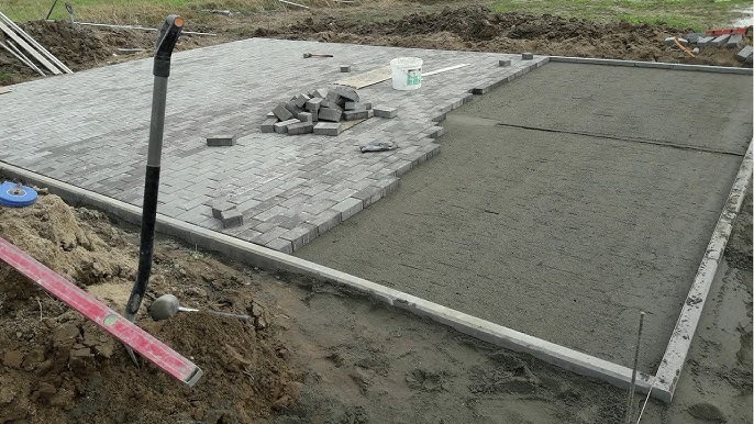 Etap układania szarej kostki brukowej na podsypce cementowej, widoczne narzędzia: łopata, poziomica, gumowy młotek i wiadro. Część powierzchni już wyłożona, część przygotowana pod kostkę, obrzeża...