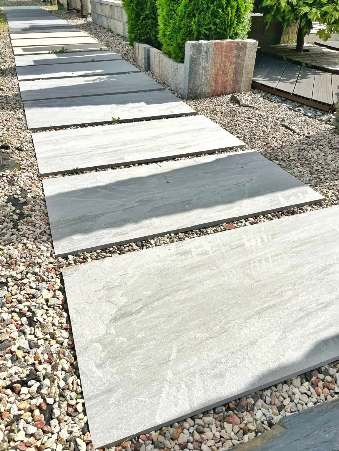 Ścieżka z dużych, prostokątnych płyt w odcieniach szarości, ułożonych w regularnych odstępach na żwirowym podłożu, prowadząca wzdłuż żywopłotu z tuj.