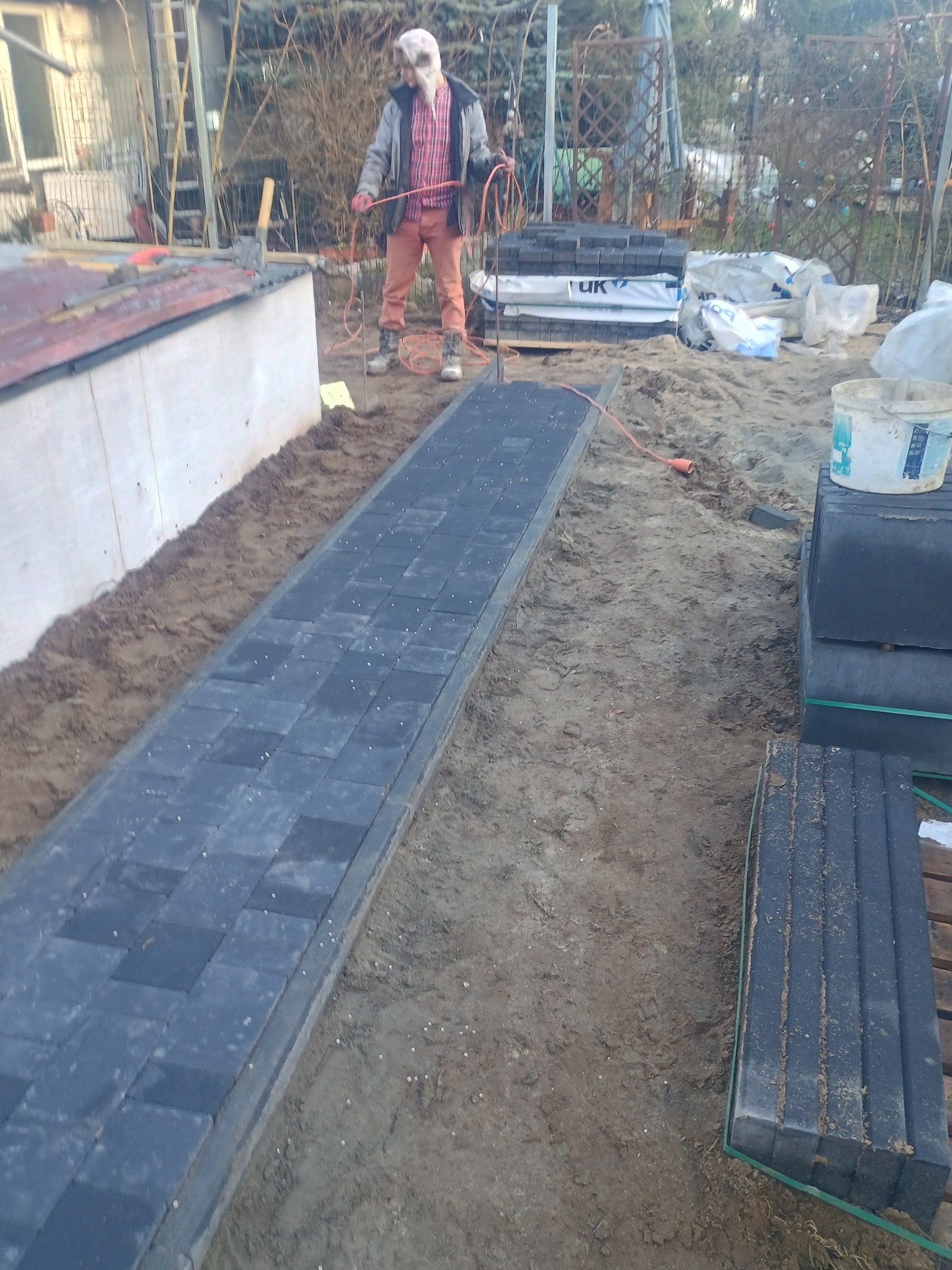 Częściowo ułożona ścieżka z ciemnej kostki brukowej z obrzeżami, obok stosy kostki, pracownik z kablem w tle, teren przygotowany pod dalsze prace.