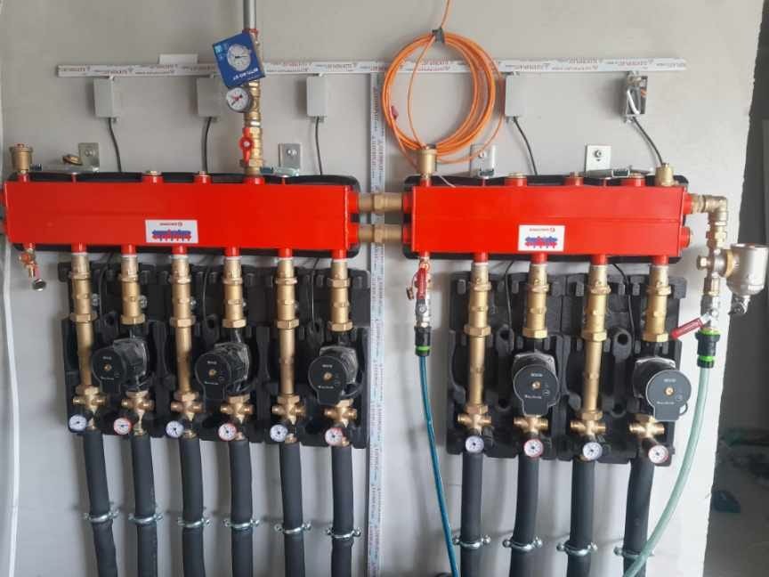 Rozdzielacz ogrzewania podłogowego z pompami obiegowymi, manometrami i rurami. Widoczne elementy instalacji zamontowane na ścianie w surowym stanie.