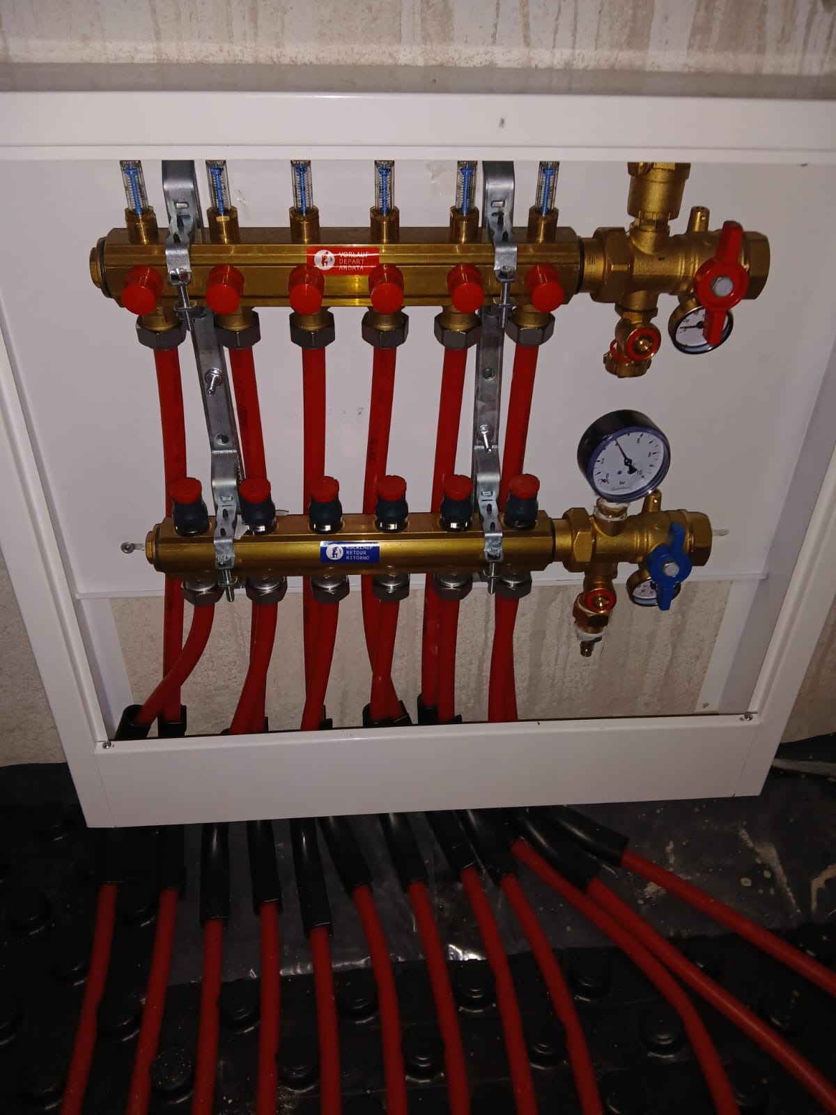 Rozdzielacz ogrzewania podłogowego w białej szafce, z czerwonymi rurami i manometrem. Widoczne elementy regulacyjne i przyłącza. Instalacja w trakcie montażu.