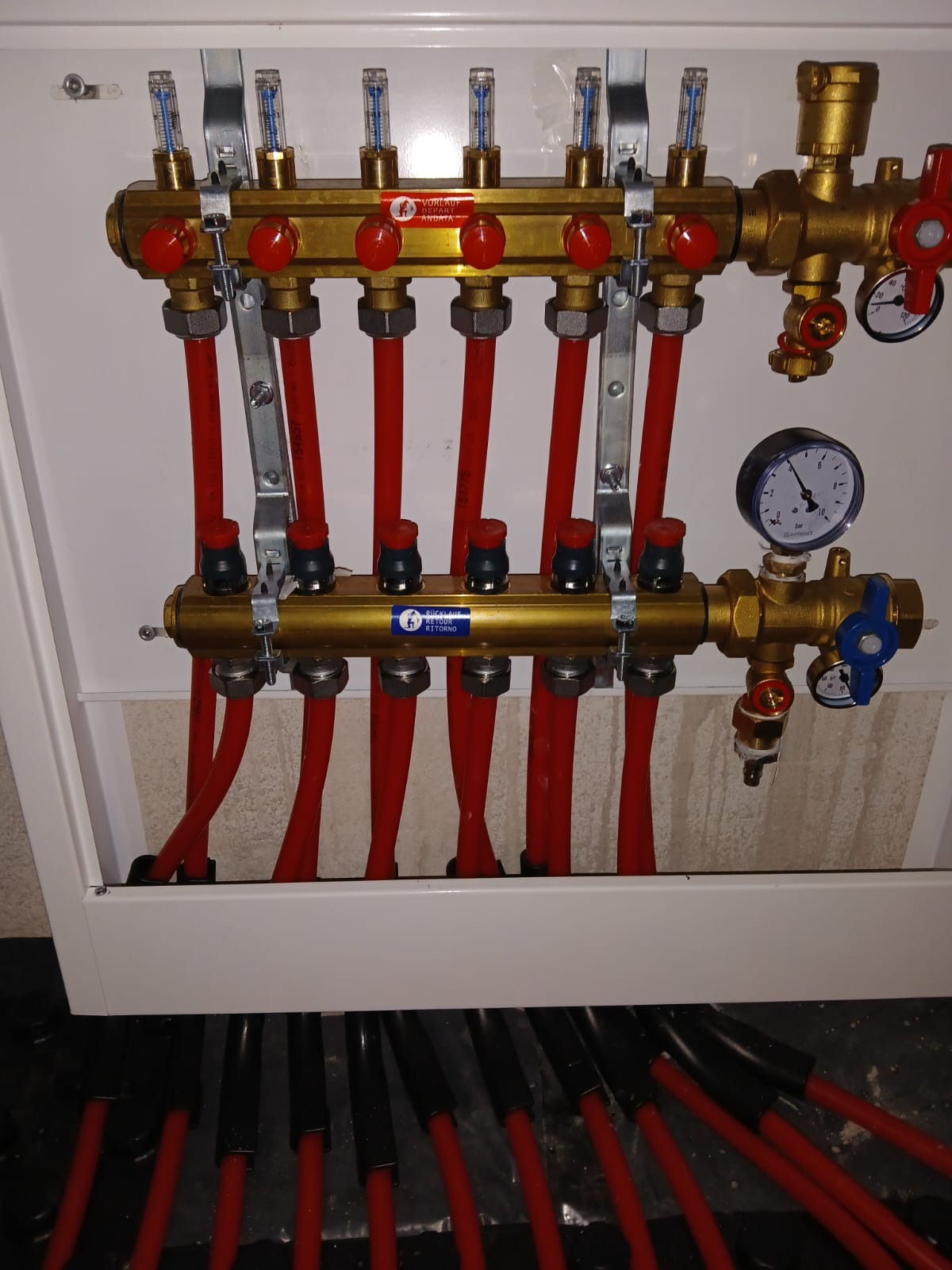 Rozdzielacz ogrzewania podłogowego z czerwonymi rurami, widoczne manometry i przepływomierze, zamontowany w białej szafce. Część rur schowana pod posadzką.