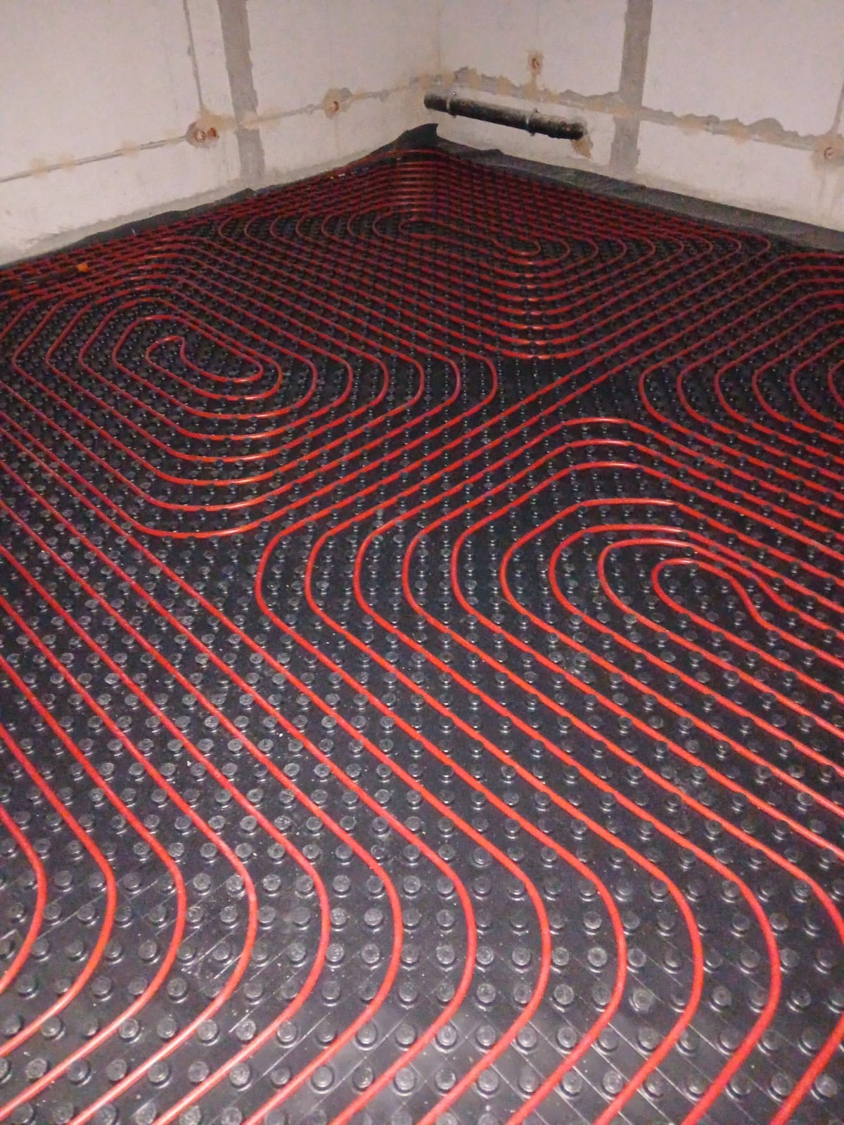 Czerwone rury ogrzewania podłogowego ułożone na czarnej macie w surowym wnętrzu. Widoczne przyłącza na ścianie.