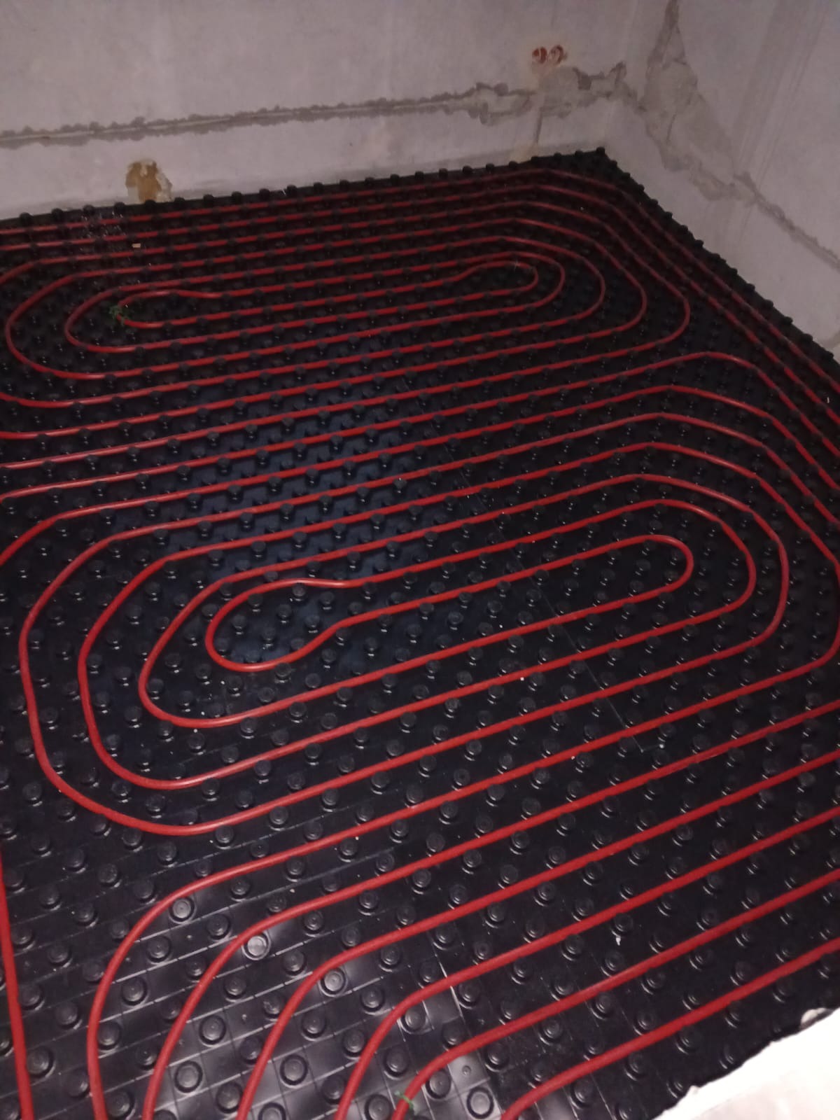 Czerwone rury ogrzewania podłogowego ułożone na czarnej macie w surowym pomieszczeniu. Widok z góry, instalacja przed wylaniem posadzki.