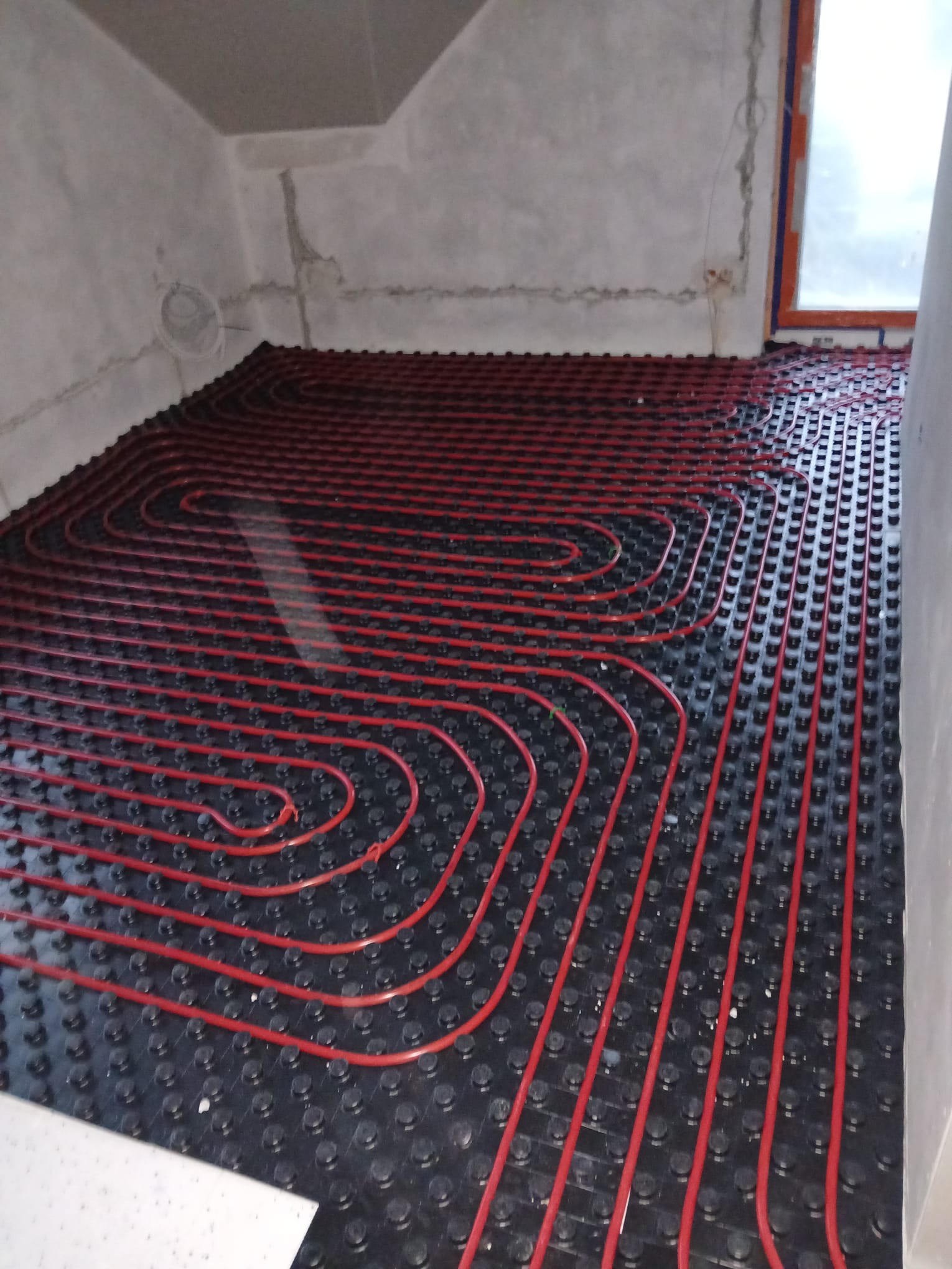 Czerwone rury ogrzewania podłogowego ułożone na czarnej macie w surowym pomieszczeniu z oknem. Widok z góry, ukośny kadr.