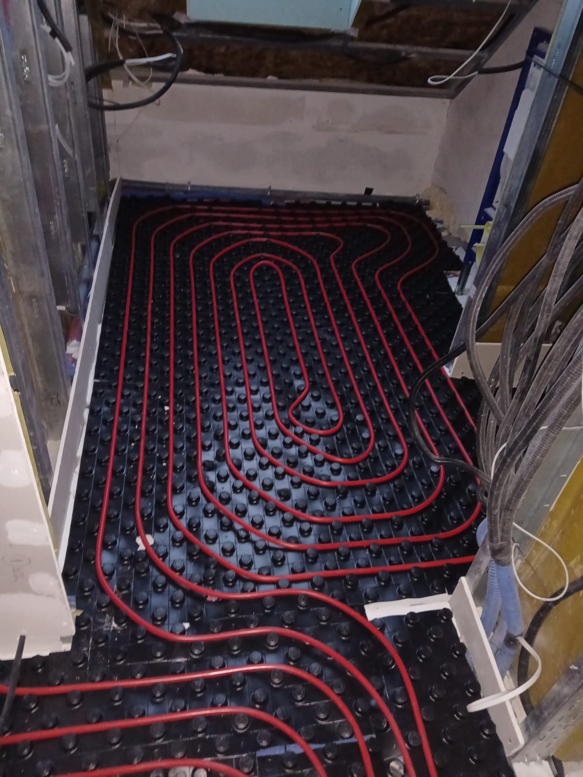 Czerwone rury ogrzewania podłogowego ułożone na czarnej macie z wypustkami, widok z góry, surowy stan wykończenia wnętrza, kable i instalacje w tle.