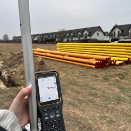 ATI-GEO ALEKSANDRA BRĘK - Pomiar geodezyjny na działce budowlanej z użyciem odbiornika GNSS, w tle rury instalacyjne i zabudowa szeregowa.
