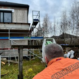 ATI-GEO ALEKSANDRA BRĘK - Człowiek w pomarańczowej kamizelce z napisem 'NANGAPARBAT EXPEDITION' obsługuje niwelator na tle budowanego domu z pustaków, widoczne rusztowania i drzewa bez liści.