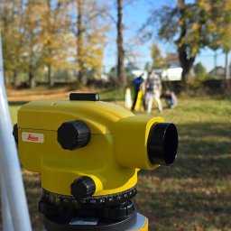 ATI-GEO ALEKSANDRA BRĘK - Żółty niwelator Leica na statywie Nivel System, w tle grupa osób podczas prac pomiarowych na zewnątrz w słoneczny dzień.