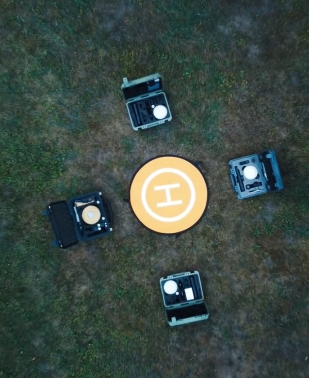 Widok z góry na cztery otwarte walizki z narzędziami pomiarowymi ułożone wokół żółtego znaku lądowiska na trawie.