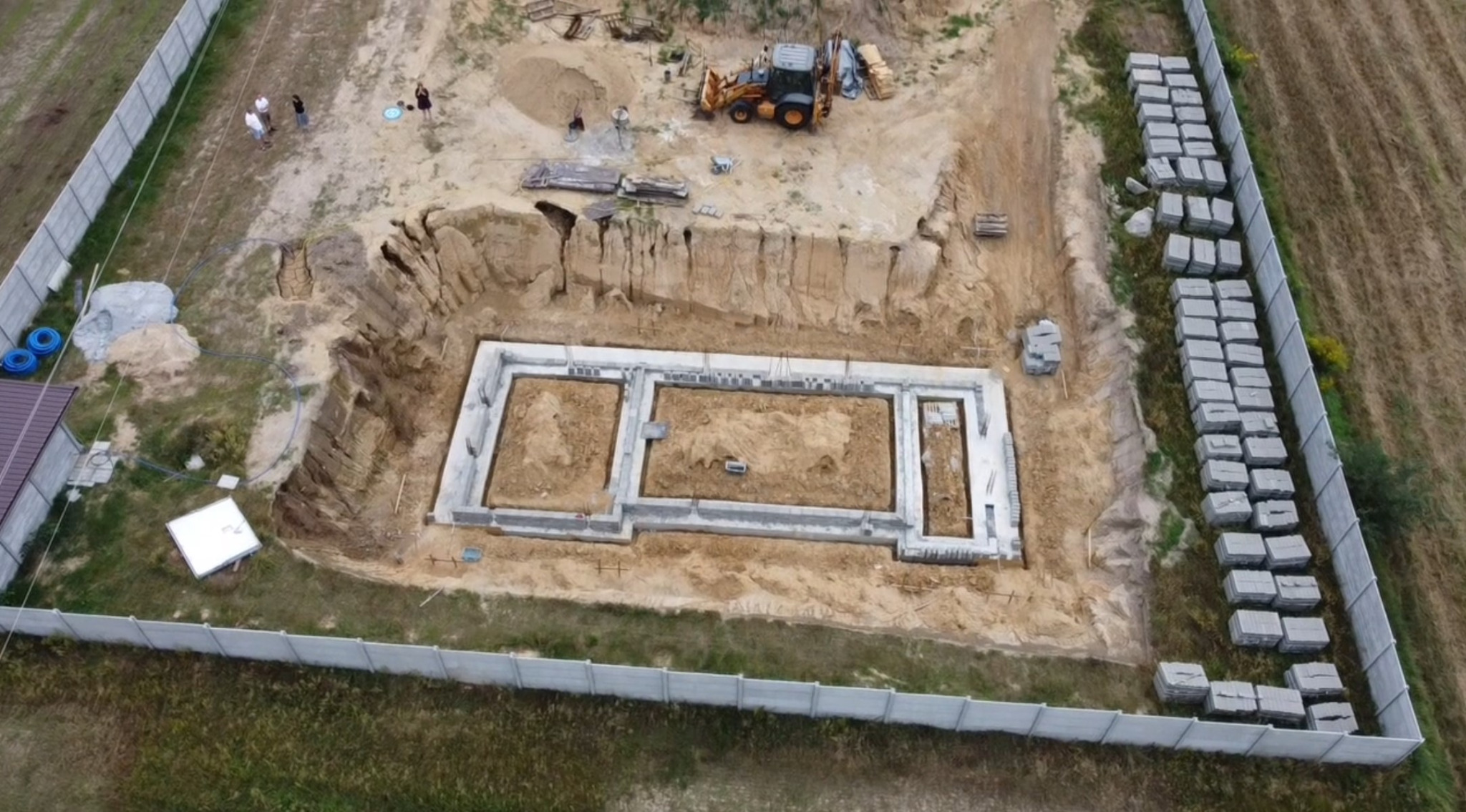 Widok z lotu ptaka na wykop pod fundamenty domu jednorodzinnego z częściowo wylanymi betonowymi fundamentami, sterty bloczków betonowych wzdłuż ogrodzenia i koparka w tle.