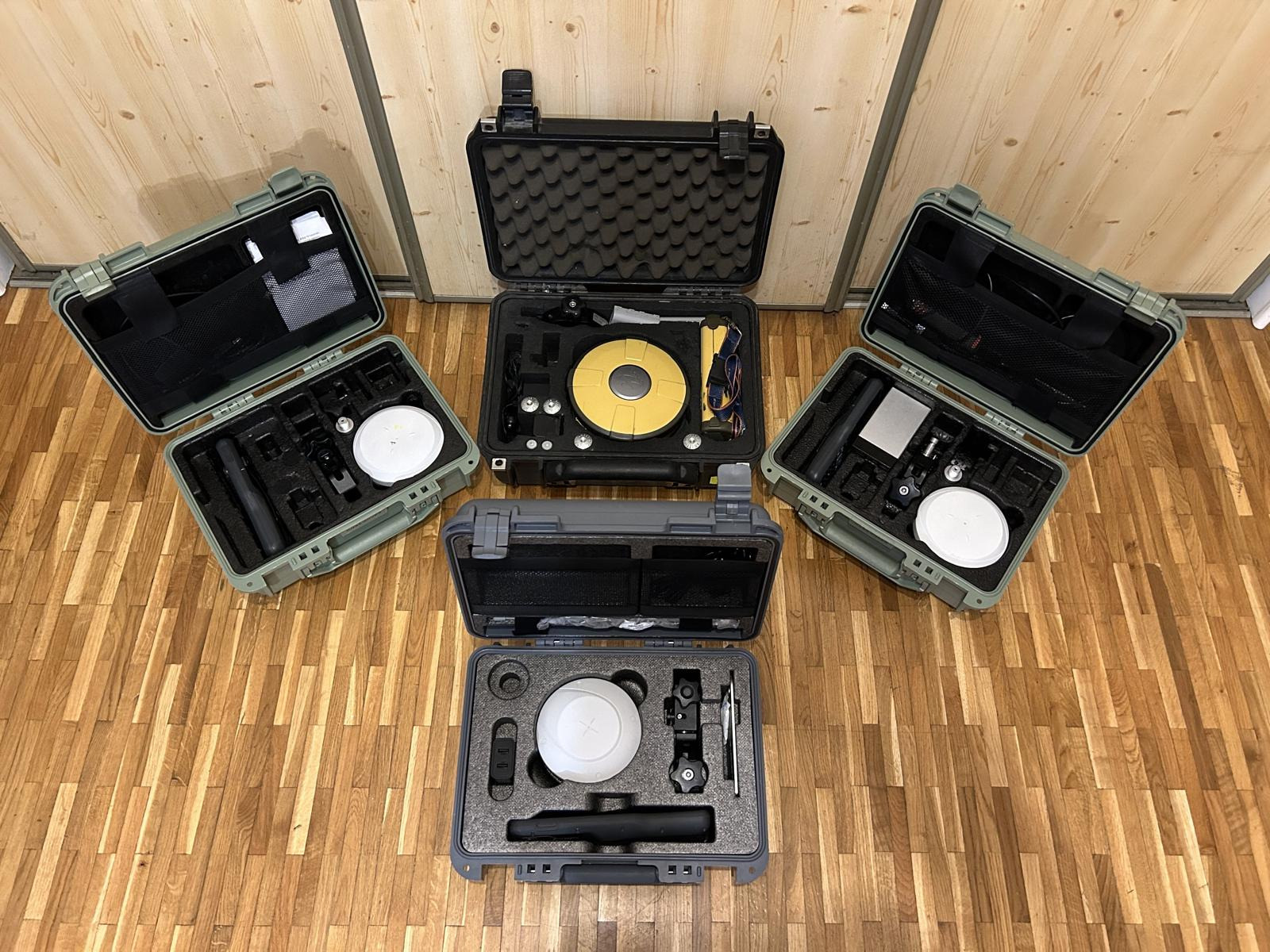 Zestaw sprzętu pomiarowego w otwartych, wyściełanych futerałach na drewnianej podłodze, widoczne anteny GPS, kontrolery i akcesoria geodezyjne.
