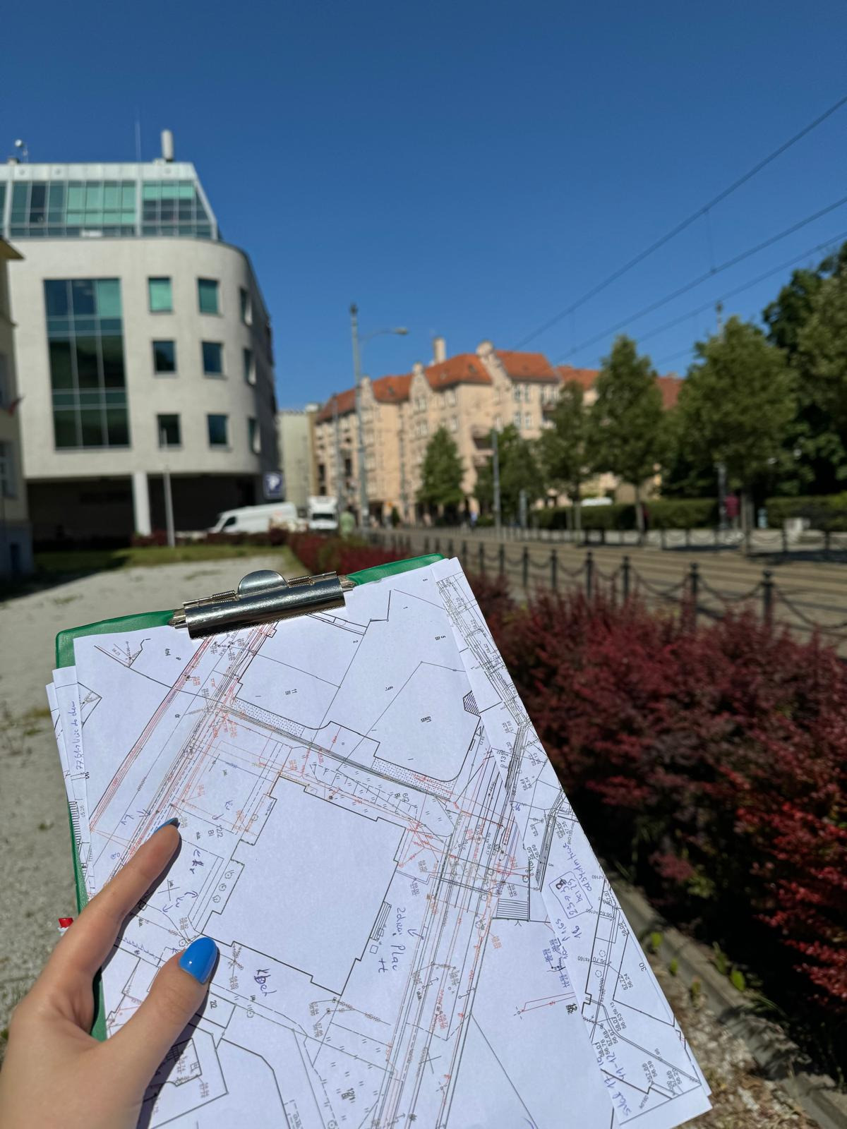 Geodeta trzymający plan zagospodarowania terenu na tle miejskiej zabudowy i czerwonych krzewów.