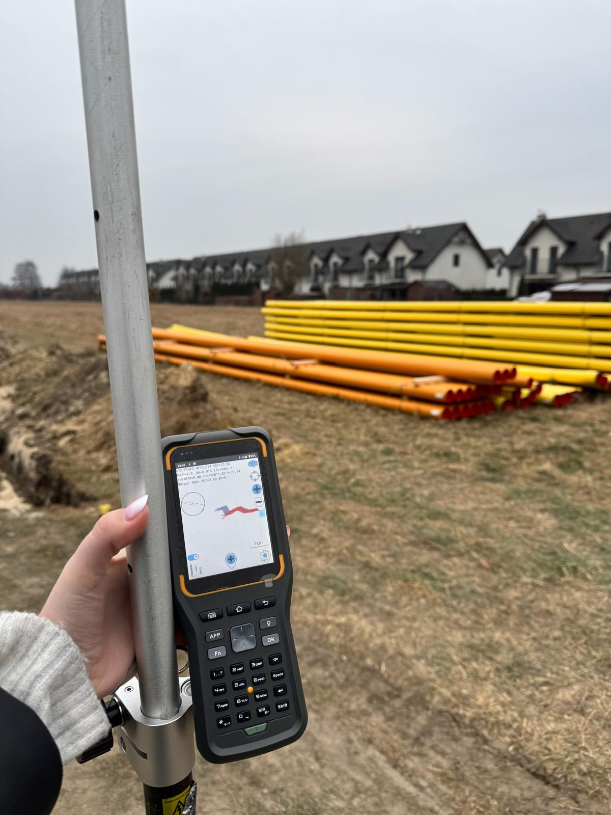 Pomiar geodezyjny na działce budowlanej z użyciem odbiornika GNSS, w tle rury instalacyjne i zabudowa szeregowa.