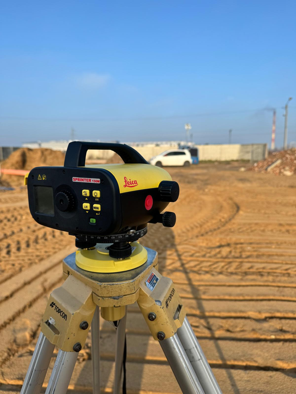 Elektroniczny niwelator Leica Sprinter 150M ustawiony na statywie Topcon na placu budowy z widocznymi śladami opon na piasku, w tle biały samochód i fragmenty konstrukcji.