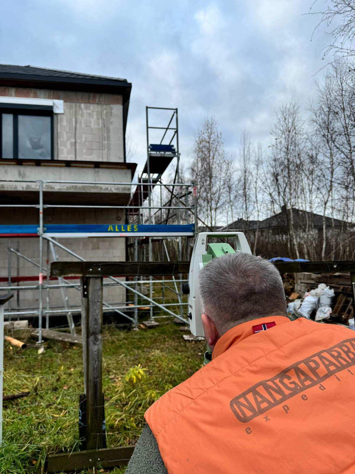 Człowiek w pomarańczowej kamizelce z napisem 'NANGAPARBAT EXPEDITION' obsługuje niwelator na tle budowanego domu z pustaków, widoczne rusztowania i drzewa bez liści.