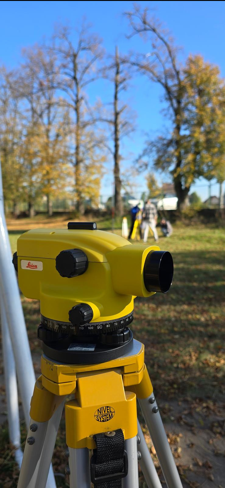 Żółty niwelator Leica na statywie Nivel System, w tle grupa osób podczas prac pomiarowych na zewnątrz w słoneczny dzień.