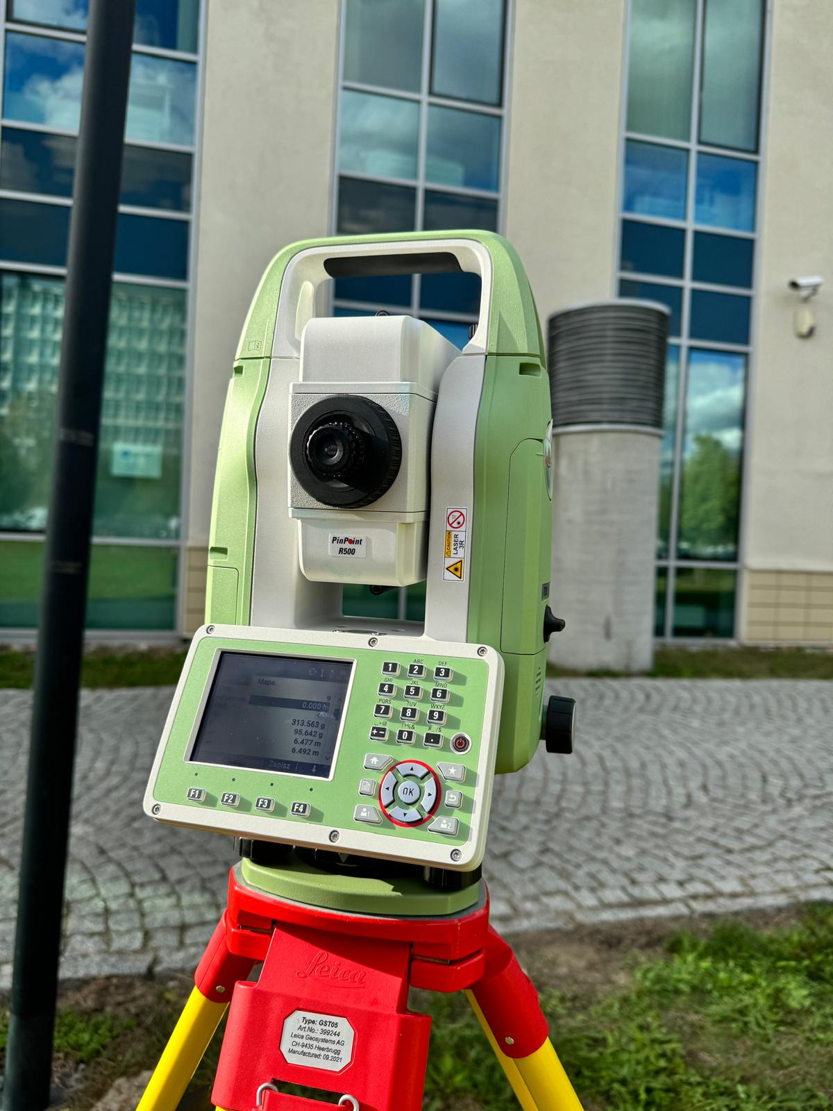 Zielono-biały teodolit Leica PinPoint R500 ustawiony na czerwono-żółtym statywie, ekran pokazuje dane pomiarowe; w tle budynek z lustrzanymi oknami i element infrastruktury.