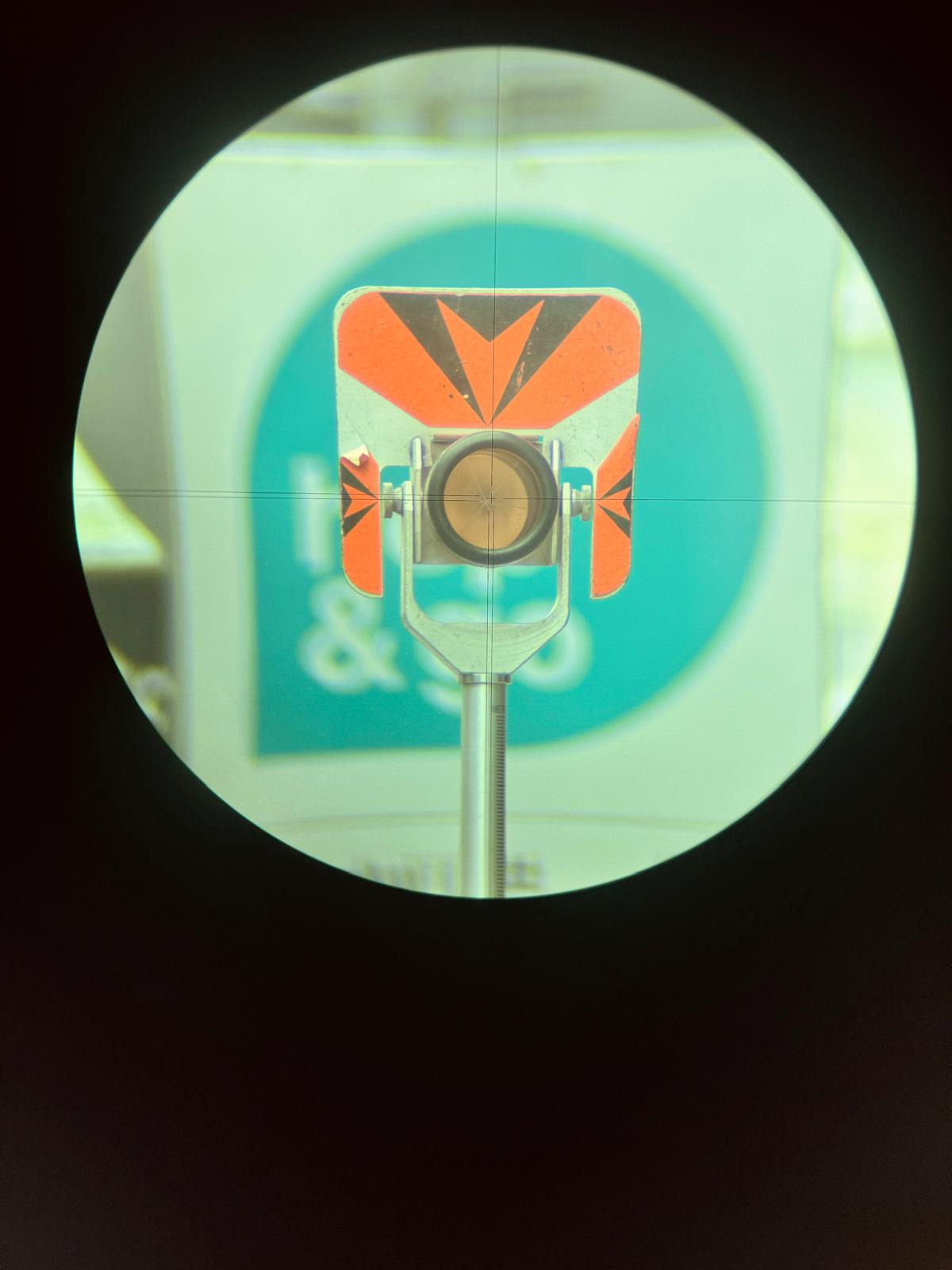 Widok przez niwelator na tarczę geodezyjną z pomarańczowymi elementami odblaskowymi i czarnymi strzałkami, ustawioną na metalowym statywie z podziałką, na rozmytym tle w kolorach seledynowo-białych.