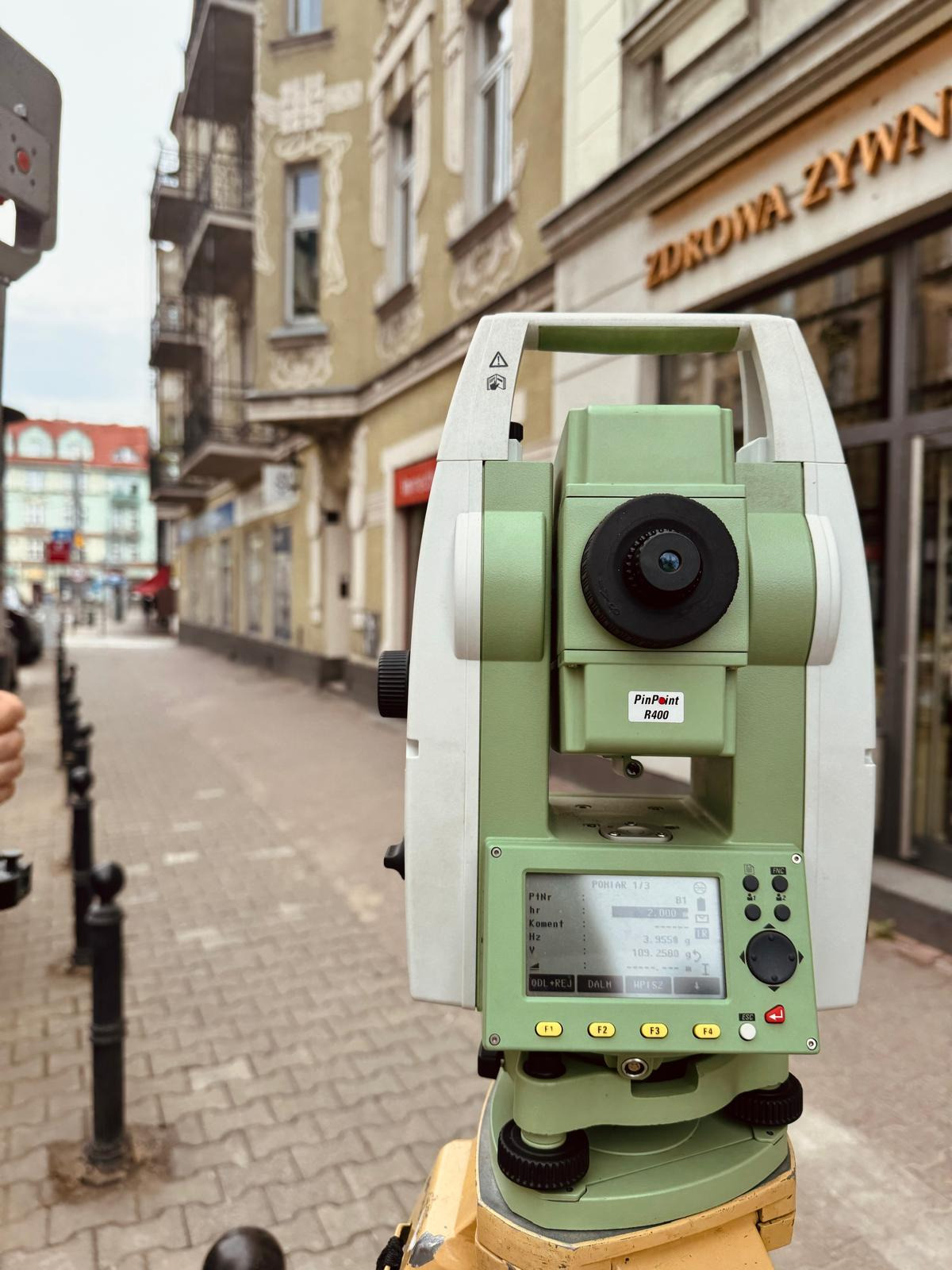 Tacheometr PinPoint R400 na tle miejskiej ulicy z budynkami, pomiar geodezyjny w trakcie realizacji.