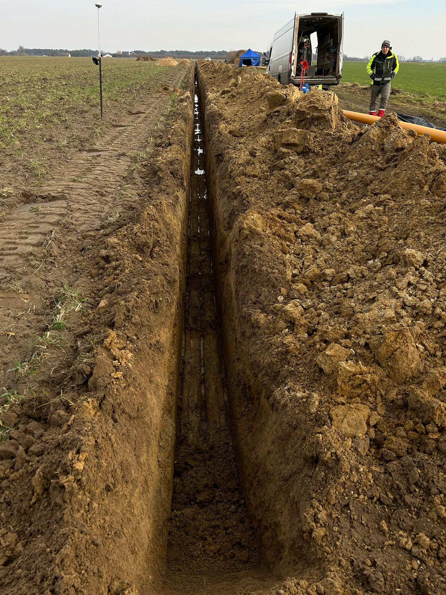 Wąski wykop w ziemi na polu, z widocznymi śladami opon i sprzętem geodezyjnym w tle, obok zaparkowany biały bus i pracownik w odblaskowej kurtce.