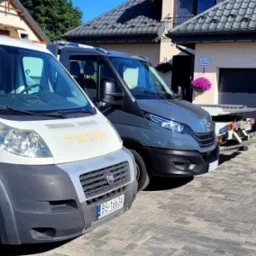 Dwa busy pomocy drogowej zaparkowane na brukowanym podjeździe przed domem z panelami słonecznymi na dachu; biały Fiat Ducato z żółtym napisem 'POMOC DROGOWA' i numerem telefonu na drzwiach, obok...