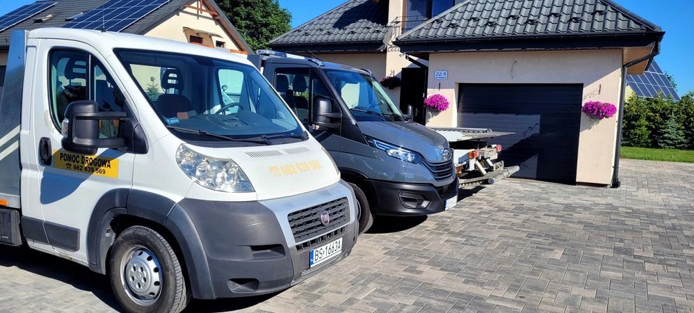 Dwa busy pomocy drogowej zaparkowane na brukowanym podjeździe przed domem z panelami słonecznymi na dachu; biały Fiat Ducato z żółtym napisem 'POMOC DROGOWA' i numerem telefonu na drzwiach, obok...