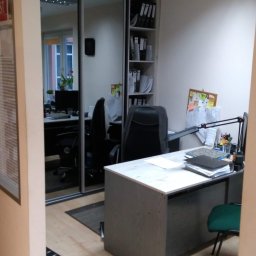 KANCELARIA DORADCY PODATKOWEGO BIURO RACHUNKOWE Paweł Kruszyński - Wnętrze biura z biurkiem, krzesłem i regałem na dokumenty. Na biurku lampka, stos dokumentów i przybory biurowe. W tle widoczne lustrzane drzwi szafy i kolejne stanowisko pracy.
